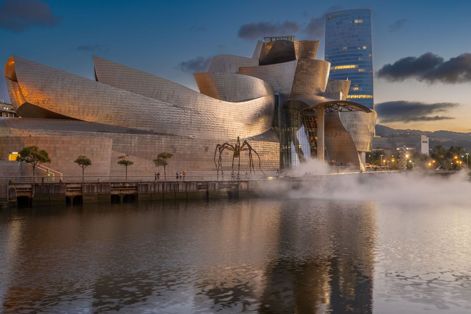 The Guggenheim Museum Bilbao reflected in water at dusk with mist and a giant spider sculpture.