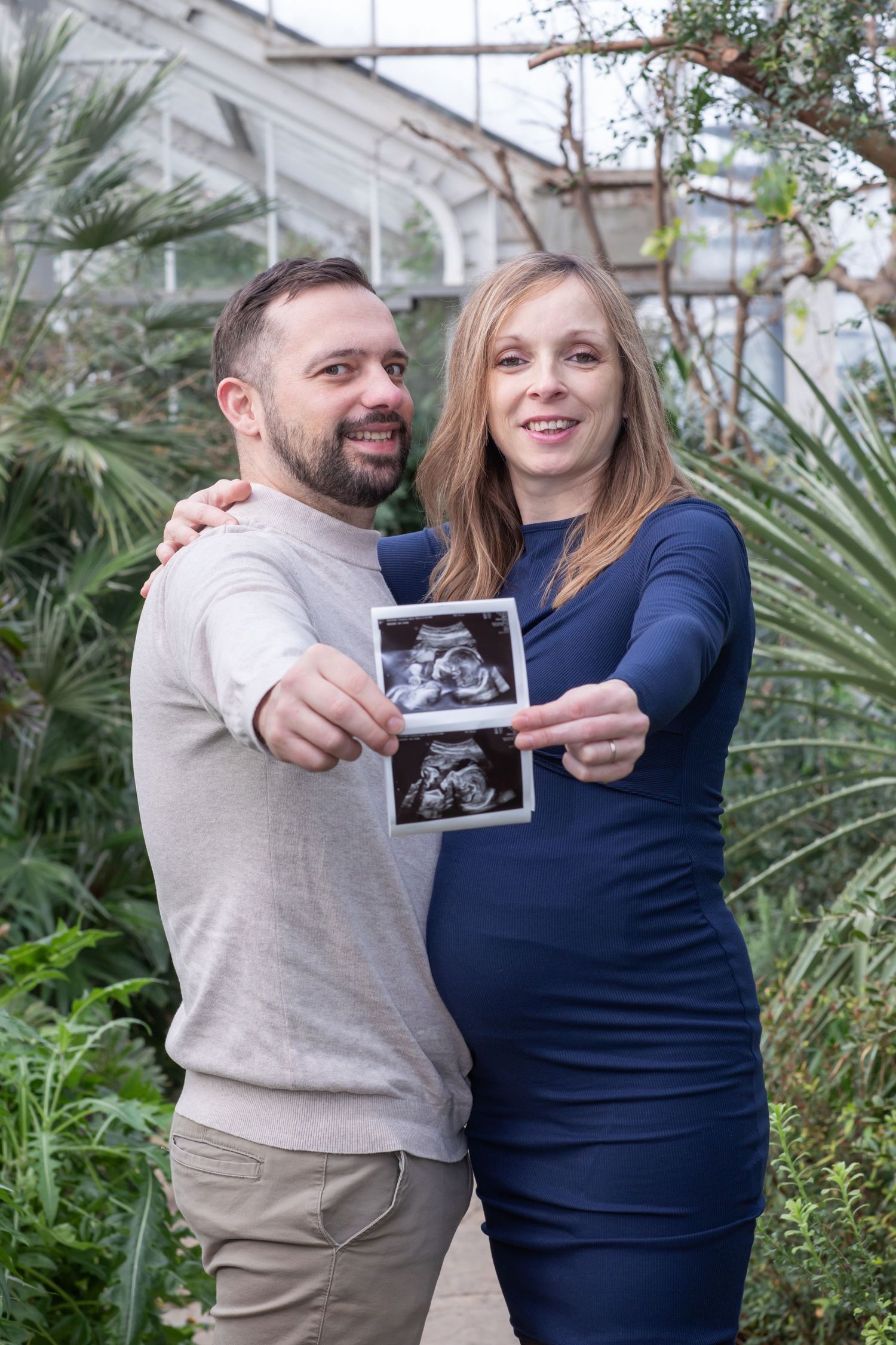 Couple in navy and beige outfits holding ultrasound photo in garden setting.