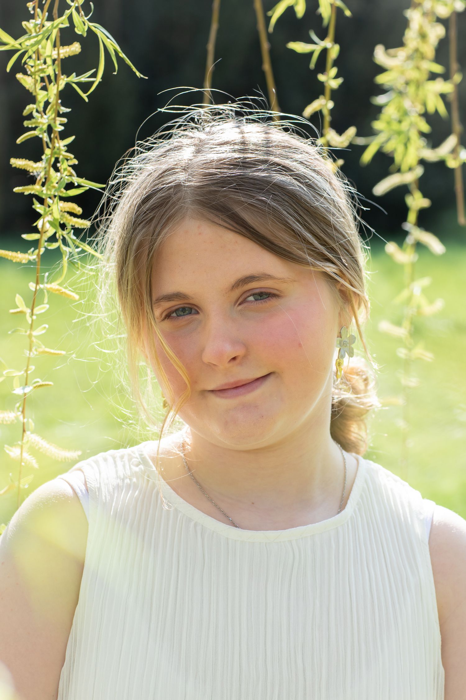 Young girl with light brown hair smiling outdoors in sunlight, wearing a white dress among green hanging branches.