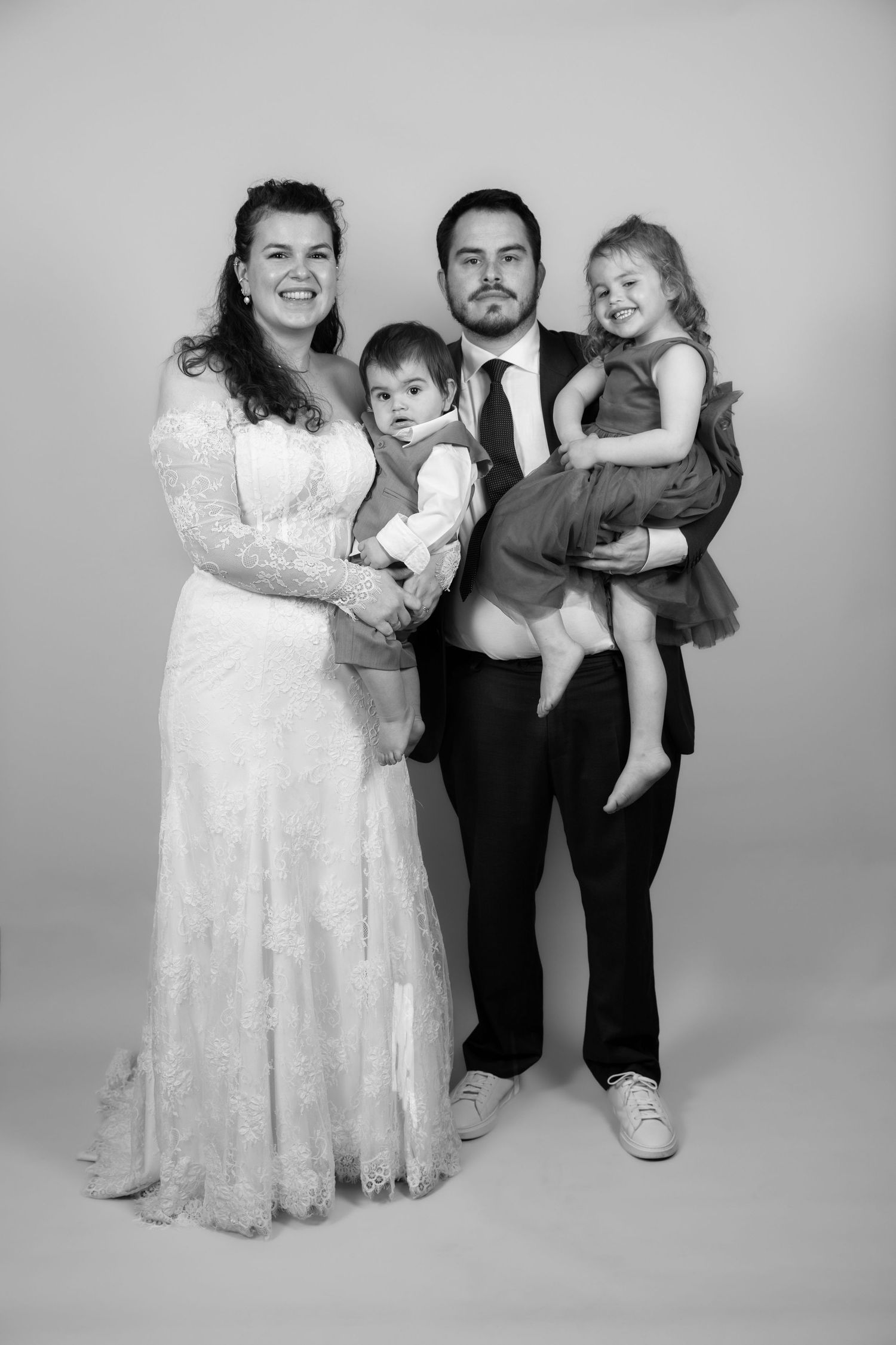 Black and white wedding photo of a couple in formal attire holding two young children, smiling in a studio setting.