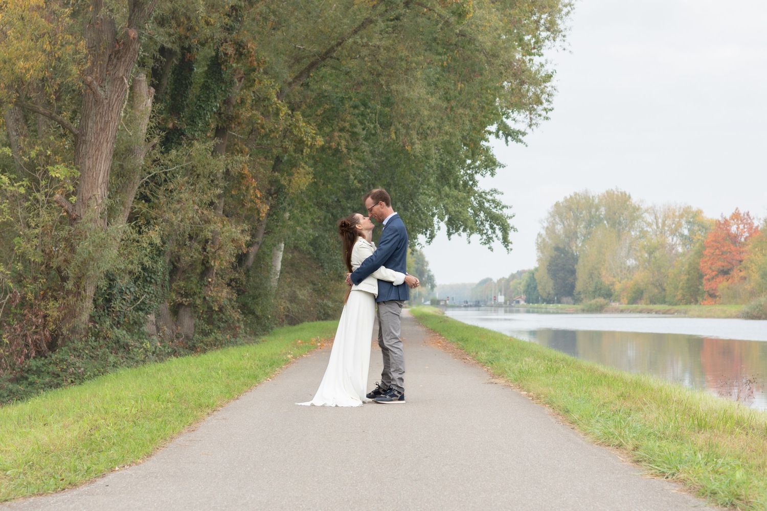A couple in wedding attire walks along a tree-lined path during autumn with fall foliage in the background.