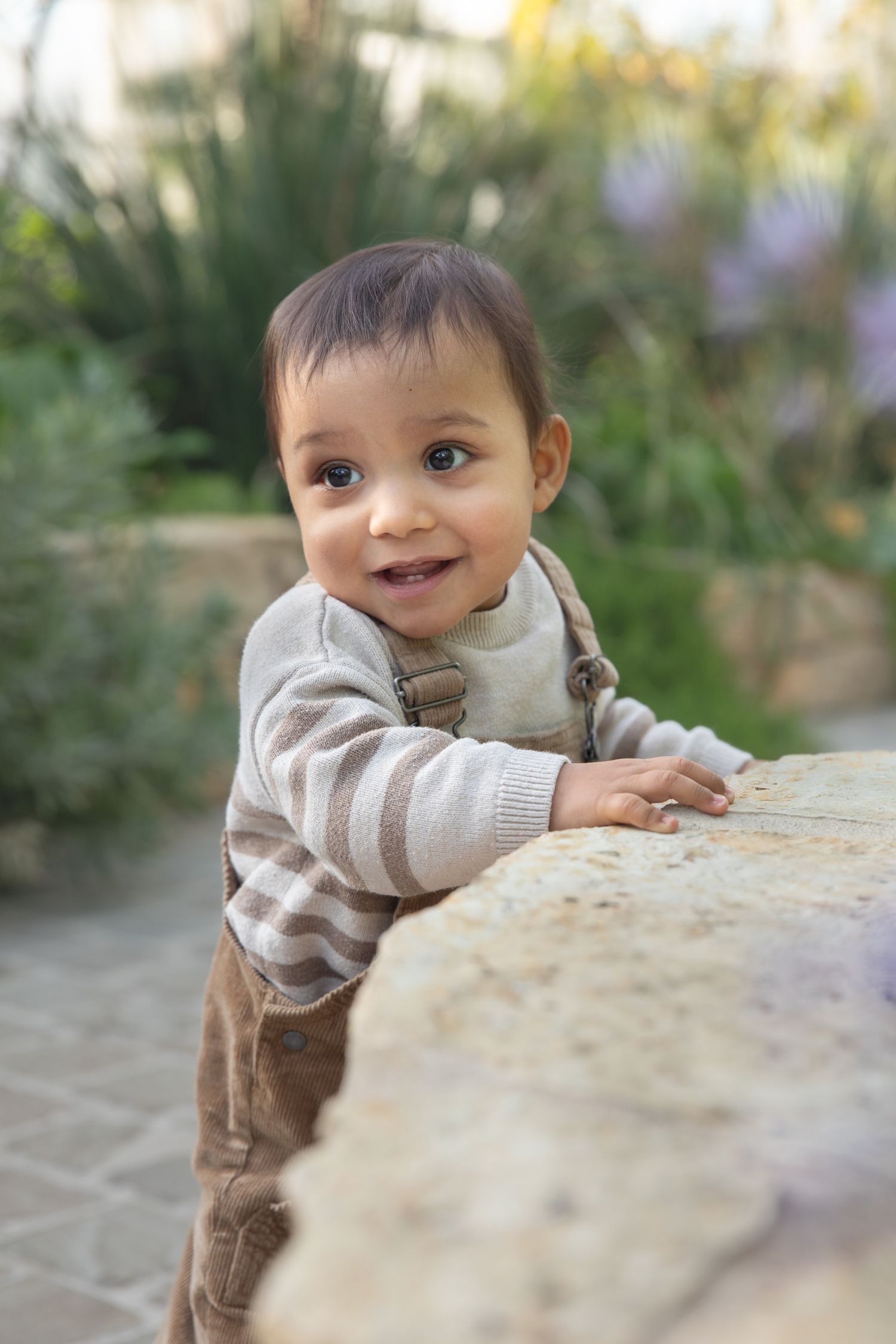 Small child in striped sweater and brown pants leans over stone surface in outdoor garden setting.