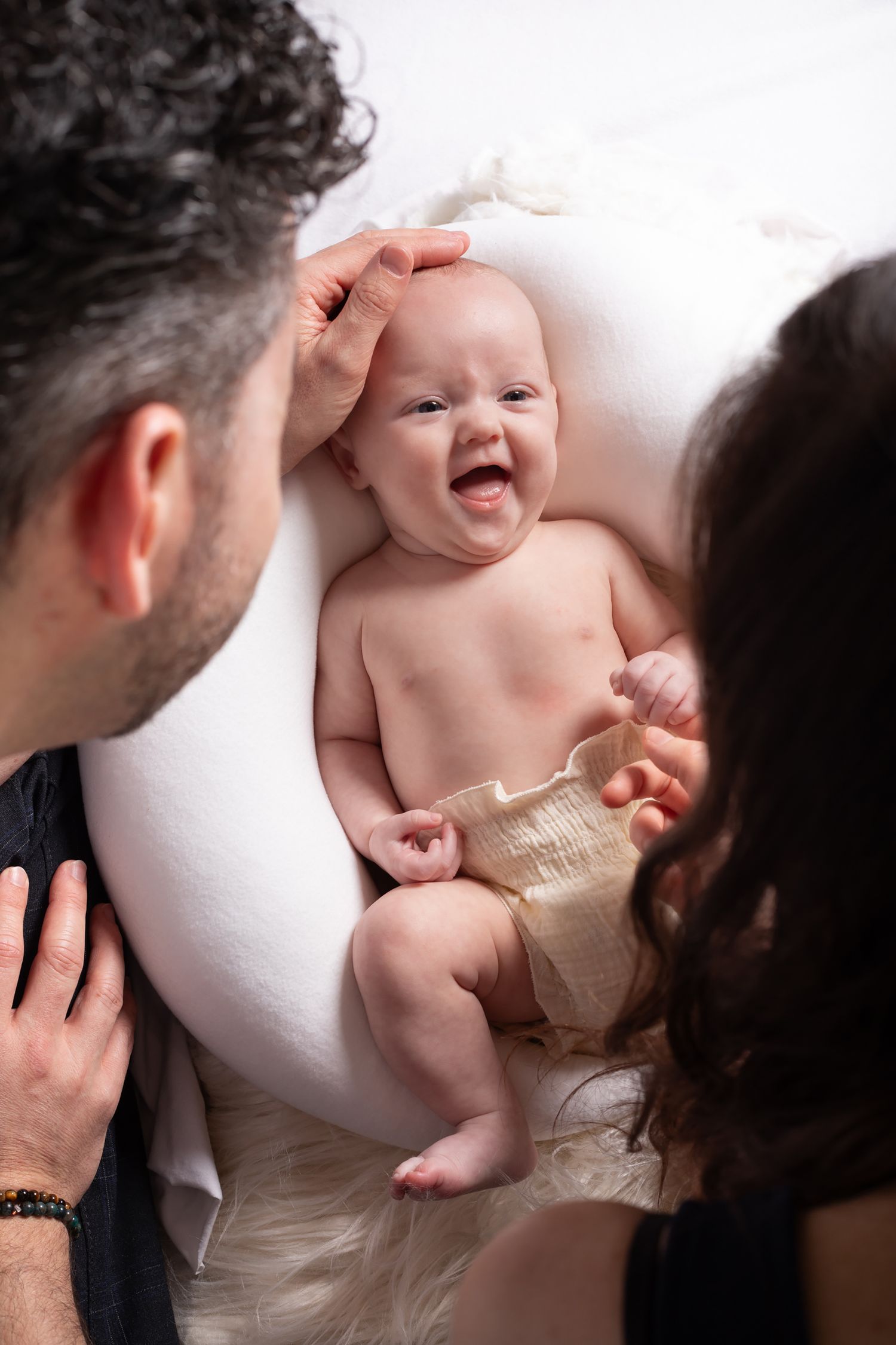 A joyful baby laughs while laying on a white surface as two adults lean in from the sides.