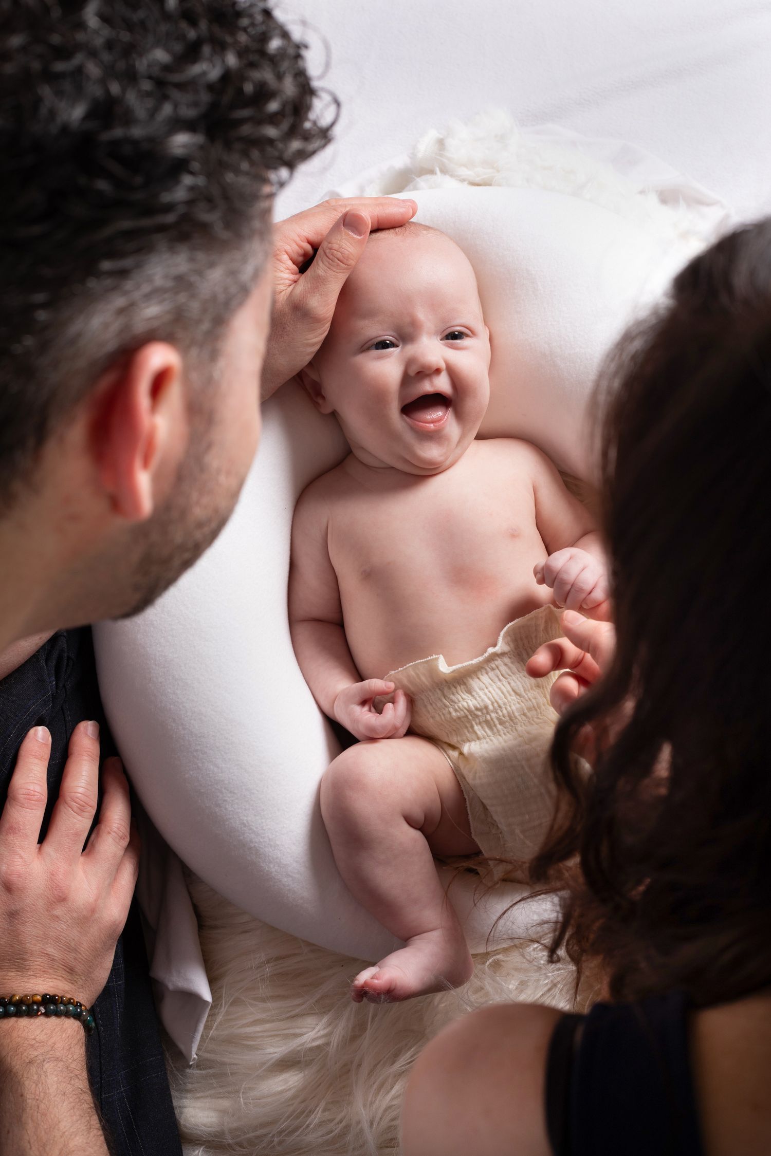 A happy baby lies on white bedding while being lovingly attended to by two adults.