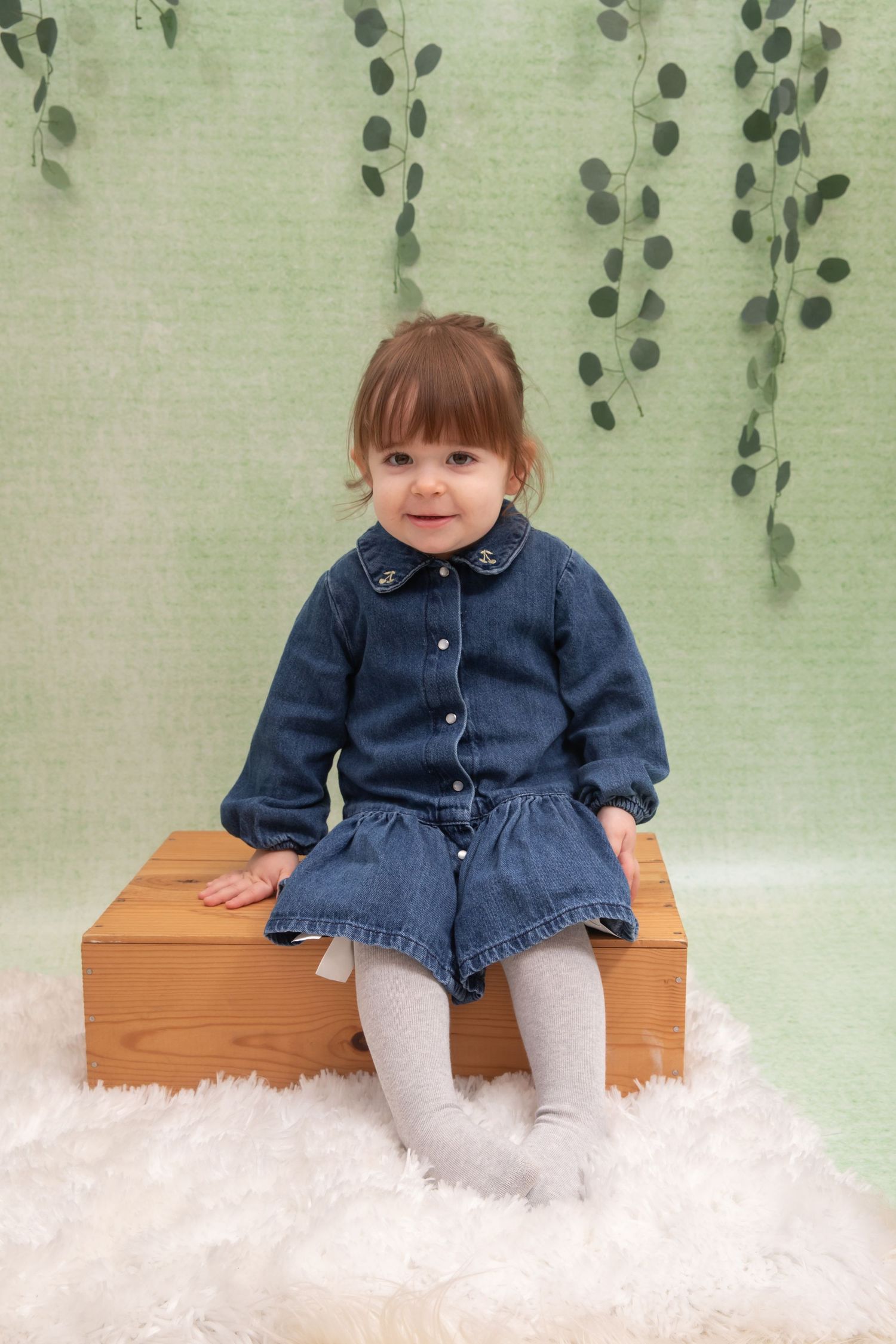 Toddler girl in denim outfit sitting on wooden box with white fur rug and green botanical backdrop.