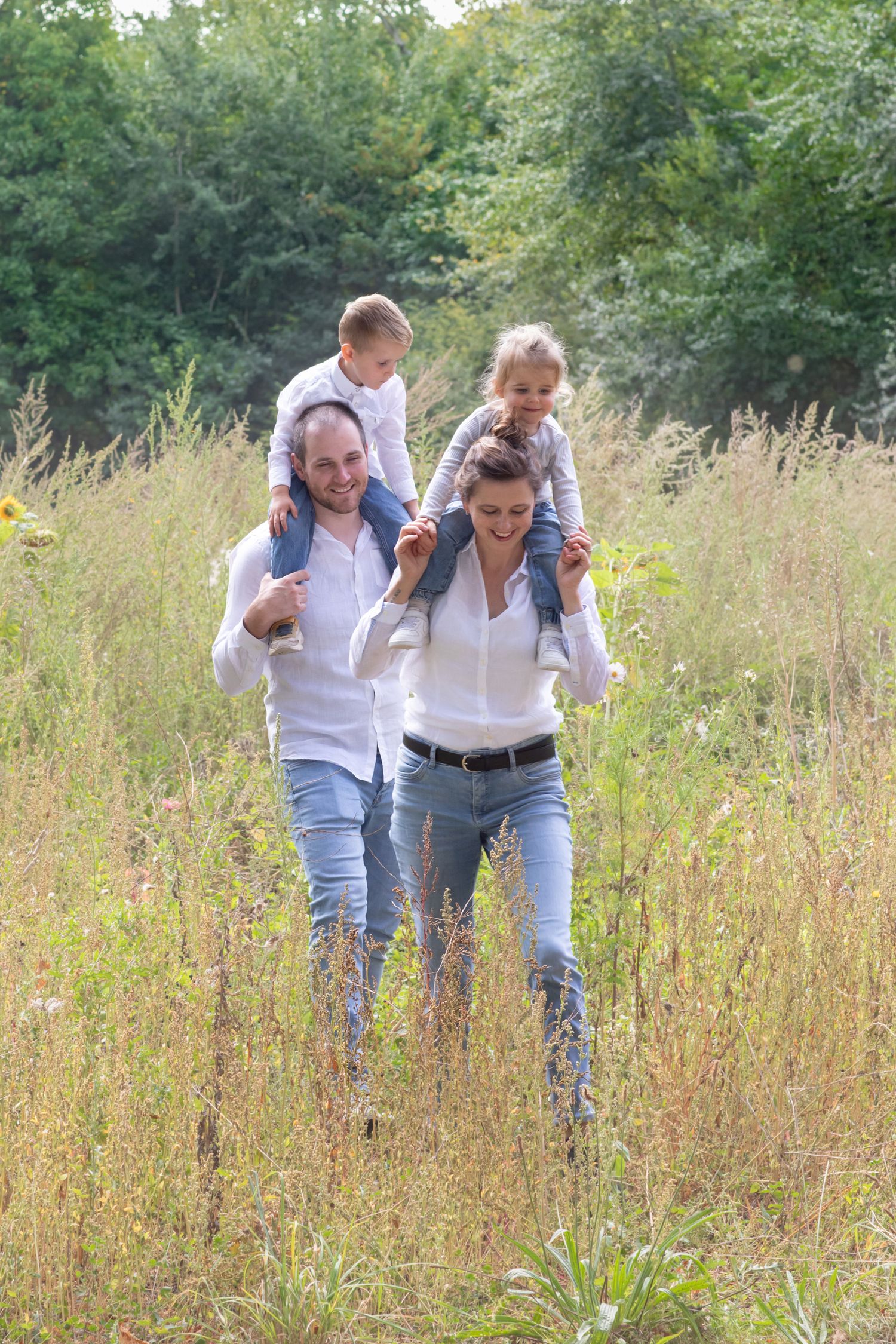 Happy family of four walking through tall grass field, parents carrying children on shoulders, wearing white shirts and jeans.