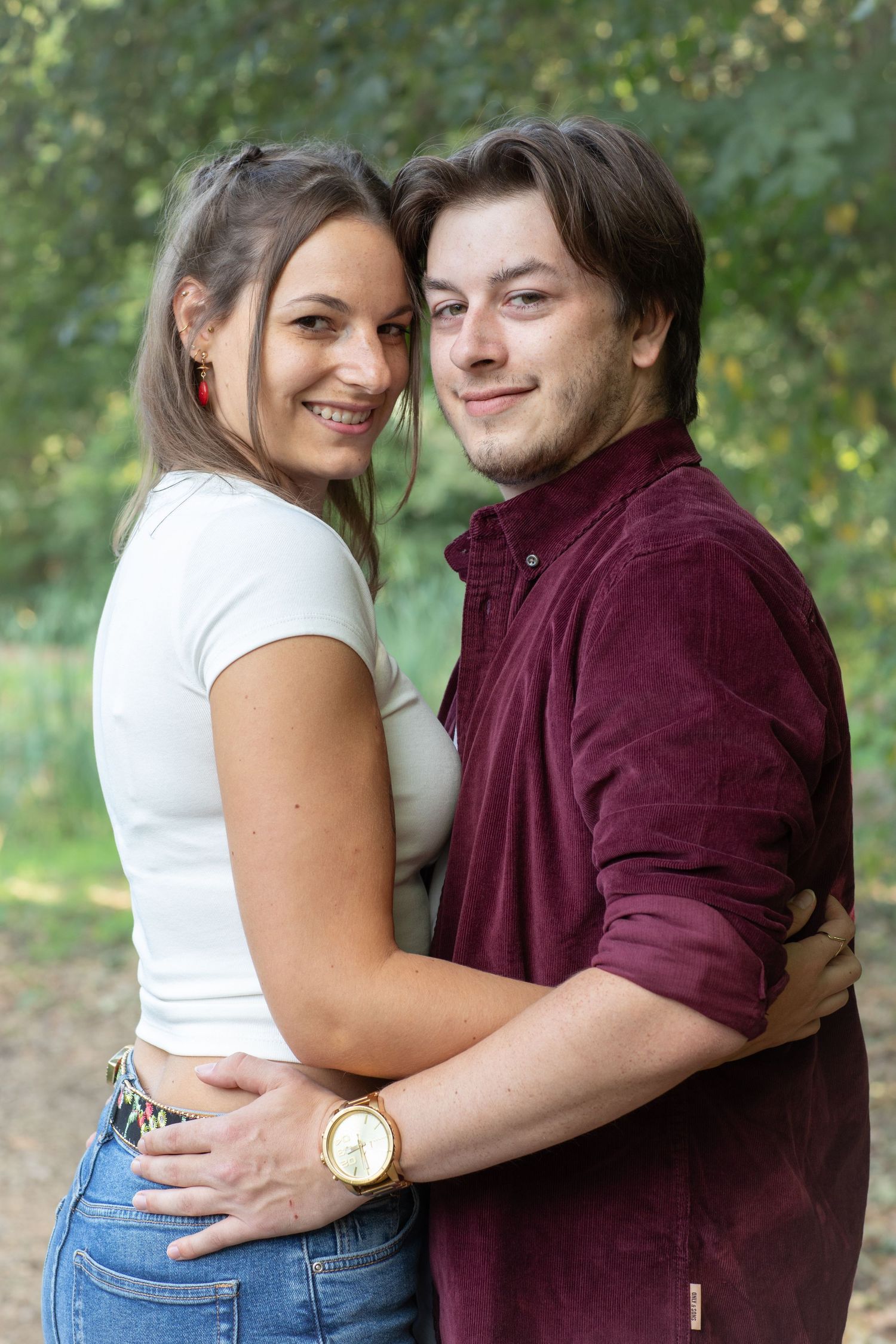 A smiling couple embracing outdoors, woman in white top and jeans, man in burgundy shirt wearing a gold watch.