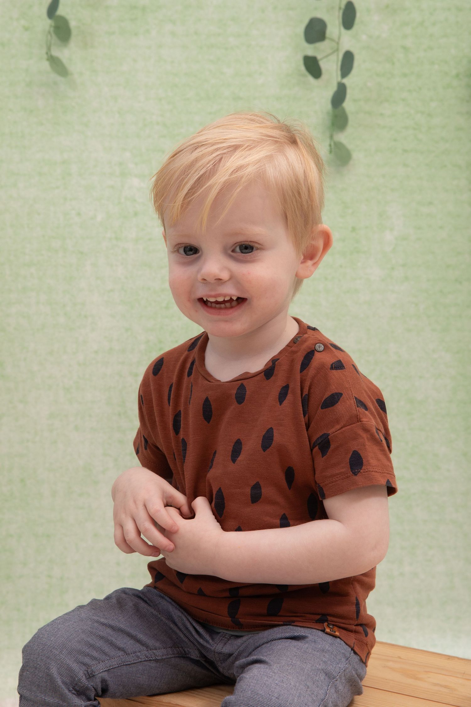 Smiling toddler boy with blonde hair wearing a brown spotted t-shirt, sitting against a green leafy background.