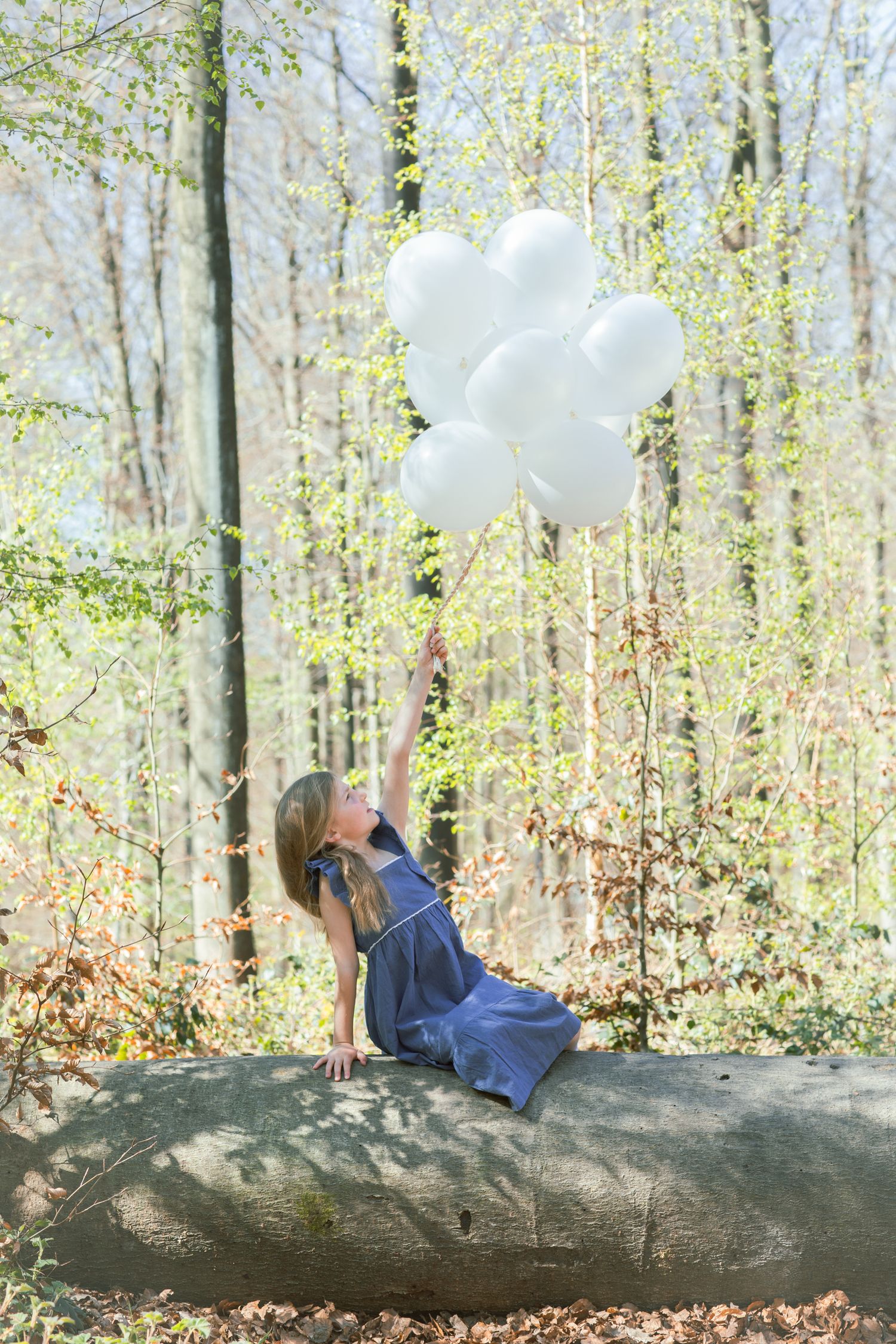 A figure in a blue dress sits on a fallen log in a spring forest while holding white balloons against a sunlit backdrop.