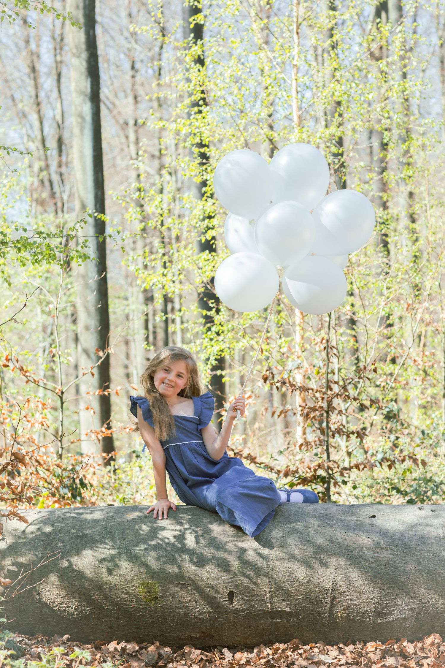 Child laying on fallen tree trunk with white balloons in spring forest setting.