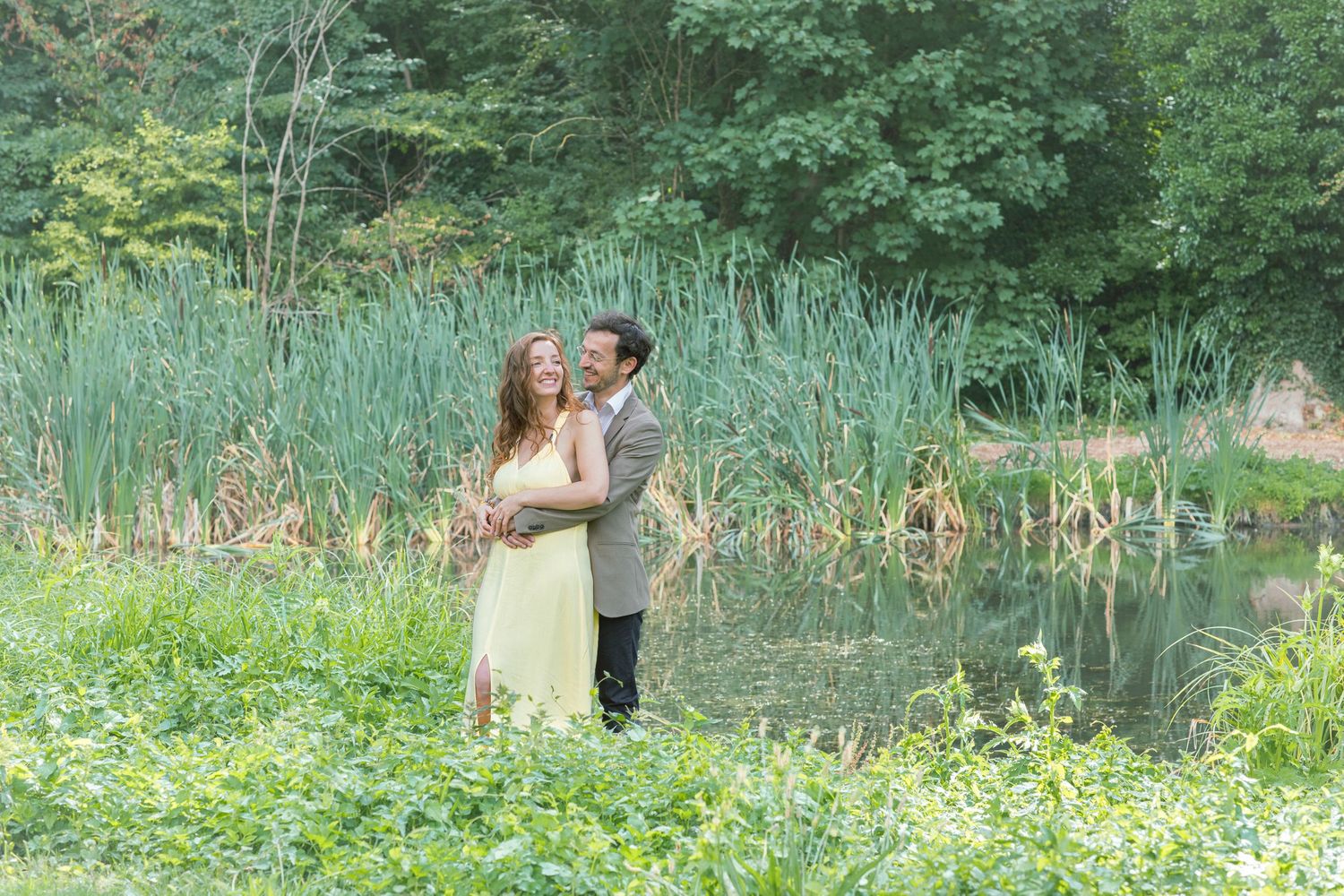 Couple embracing near a serene pond surrounded by lush green reeds and trees in a romantic outdoor setting.