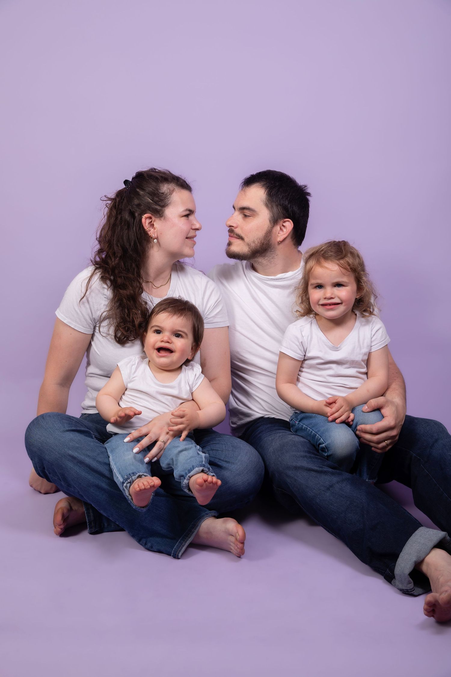 Happy family of four in white shirts and jeans sitting together on purple background in a studio photo.