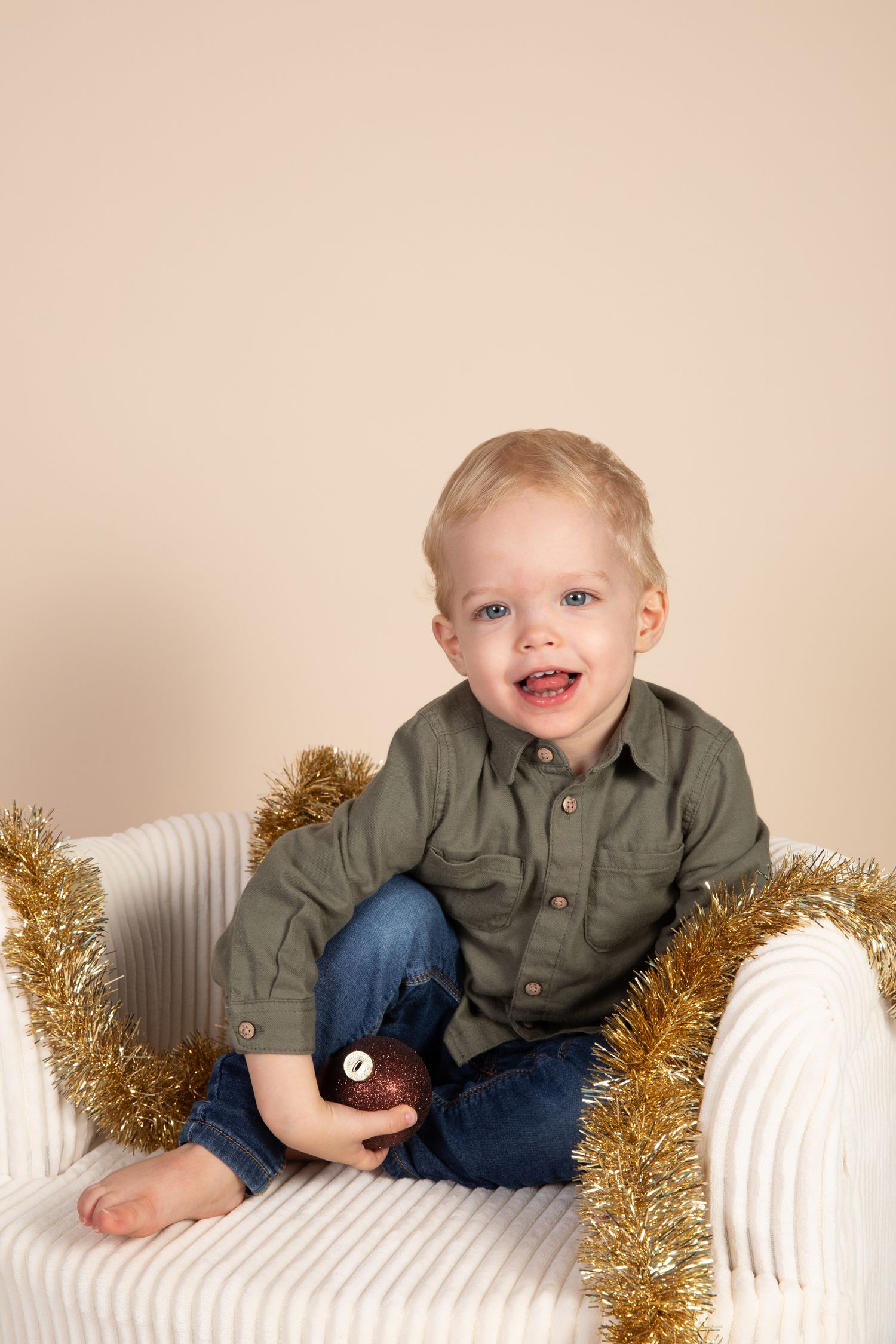 A toddler in a green button-up shirt plays with gold tinsel while sitting on a white chair against a beige background.