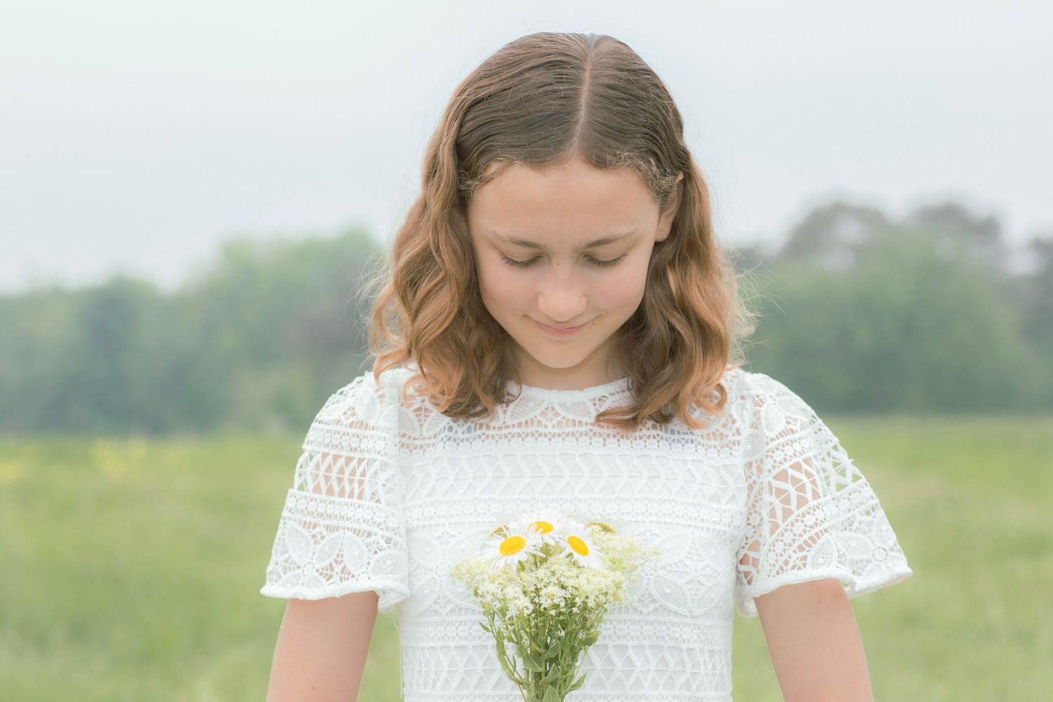 Person wearing white lace dress holding delicate wildflowers in a sunny outdoor setting.