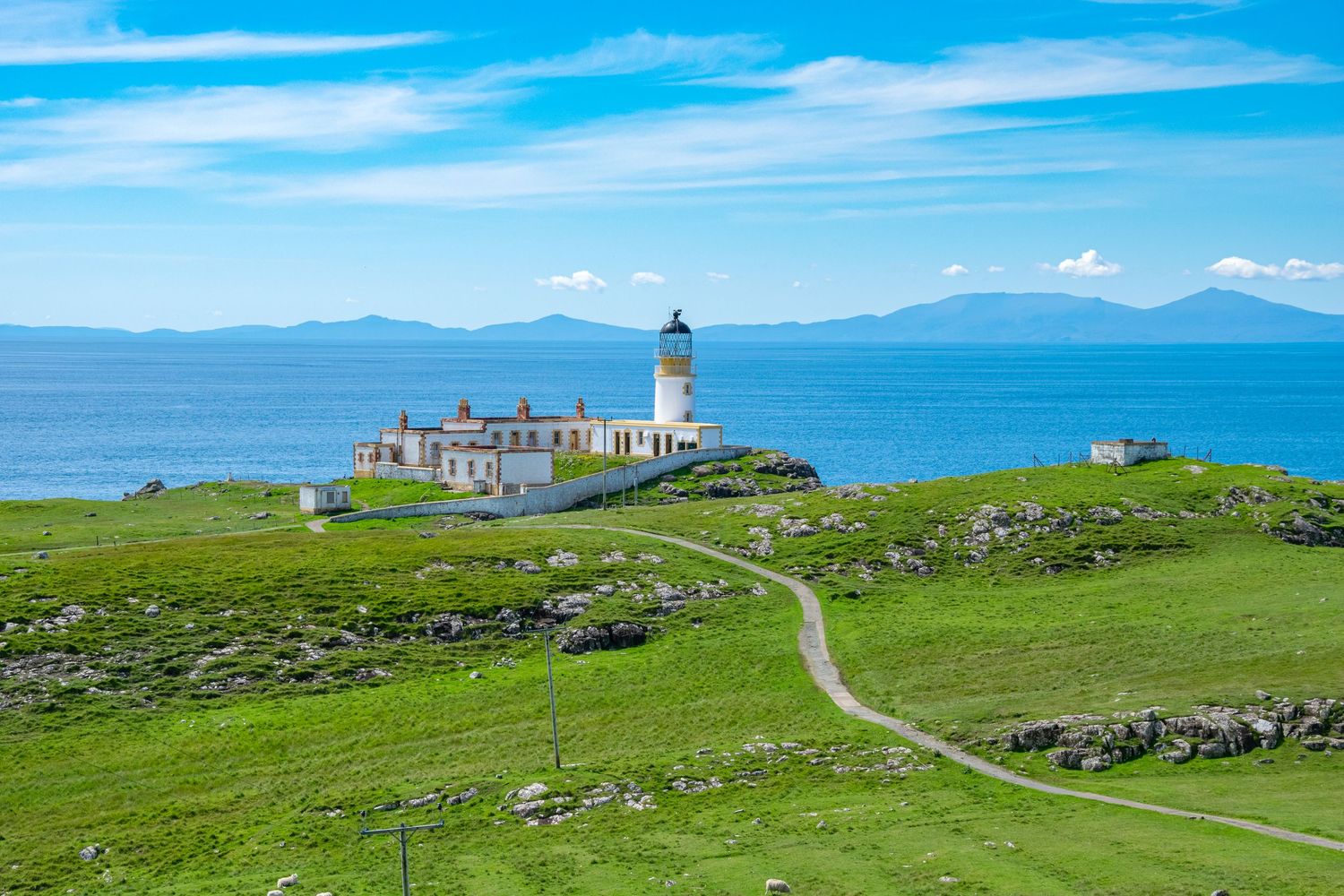 Neist Point lighthouse on green cliffs overlooking a calm blue sea with mountains in the background, Isle of Skye, Scotland.