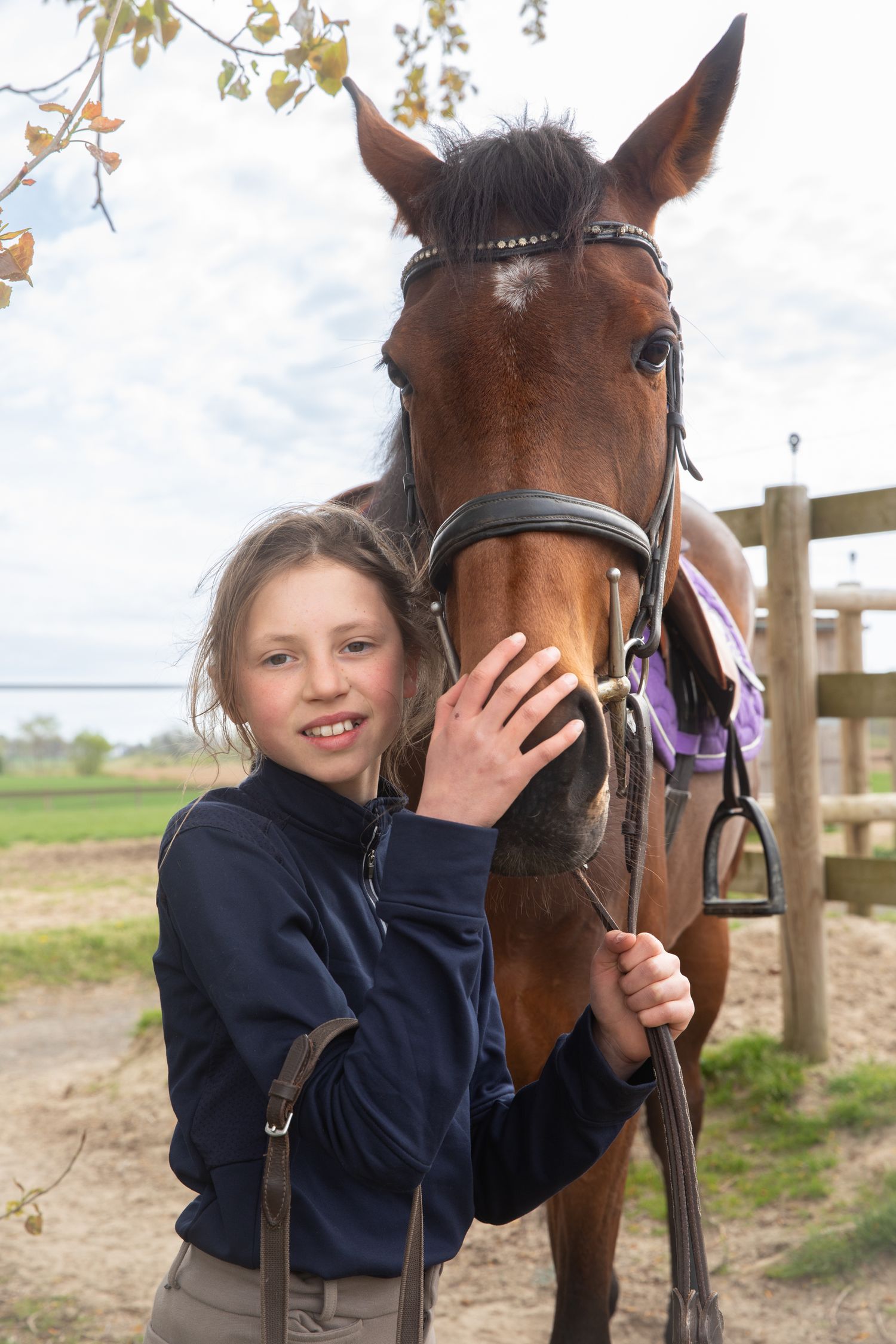Young girl smiling beside a brown horse with bridle at an equestrian facility outdoors.