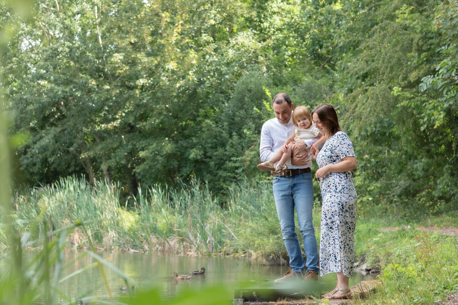 Family of three standing by a pond in a lush green forest, parents holding their baby outdoors.