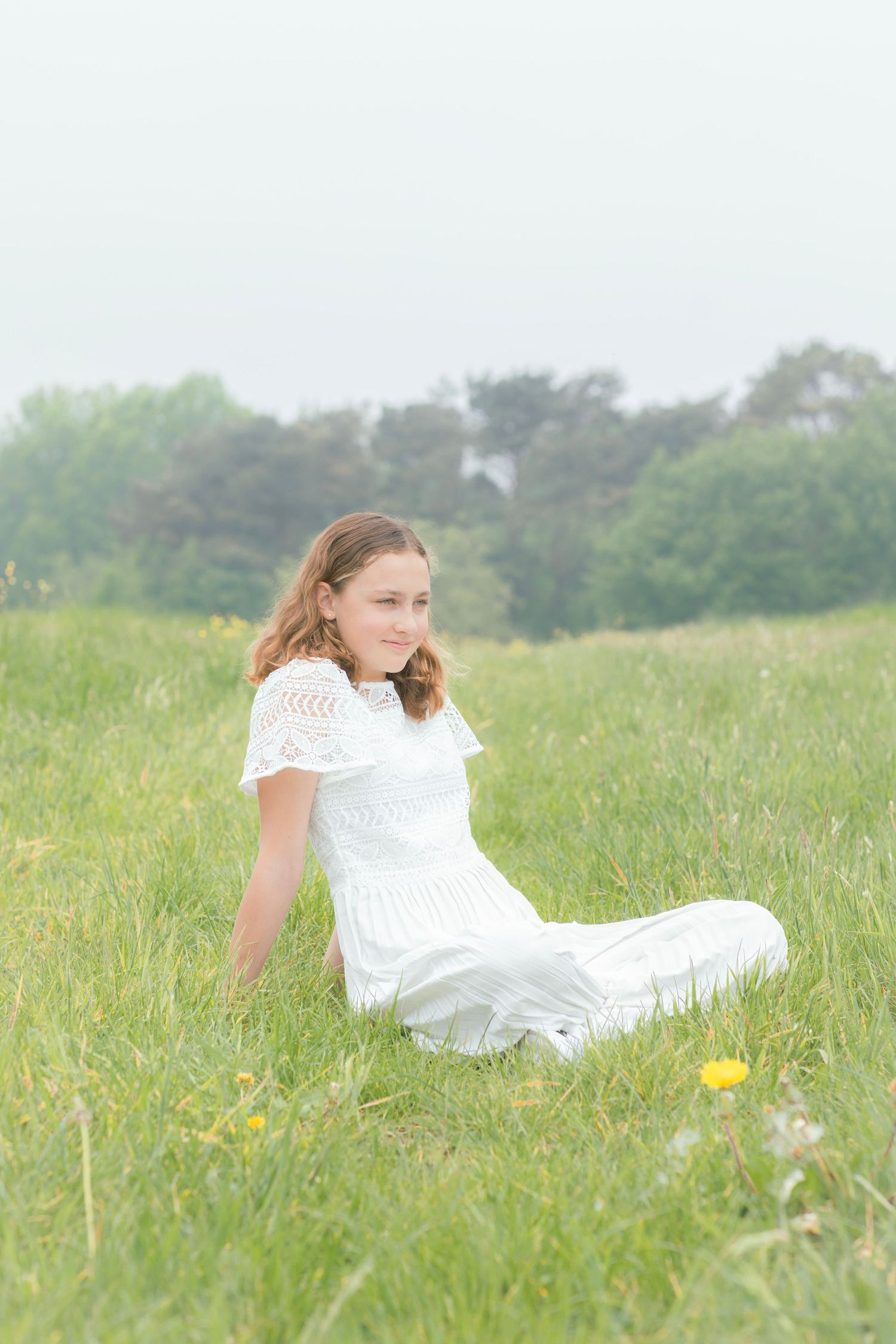 Someone in a white lace dress sits peacefully in a summer meadow with wildflowers and tall grass.