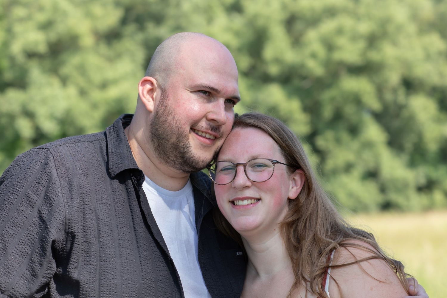 A smiling couple poses outdoors with lush green trees in the background, the man bald and the woman wearing glasses.