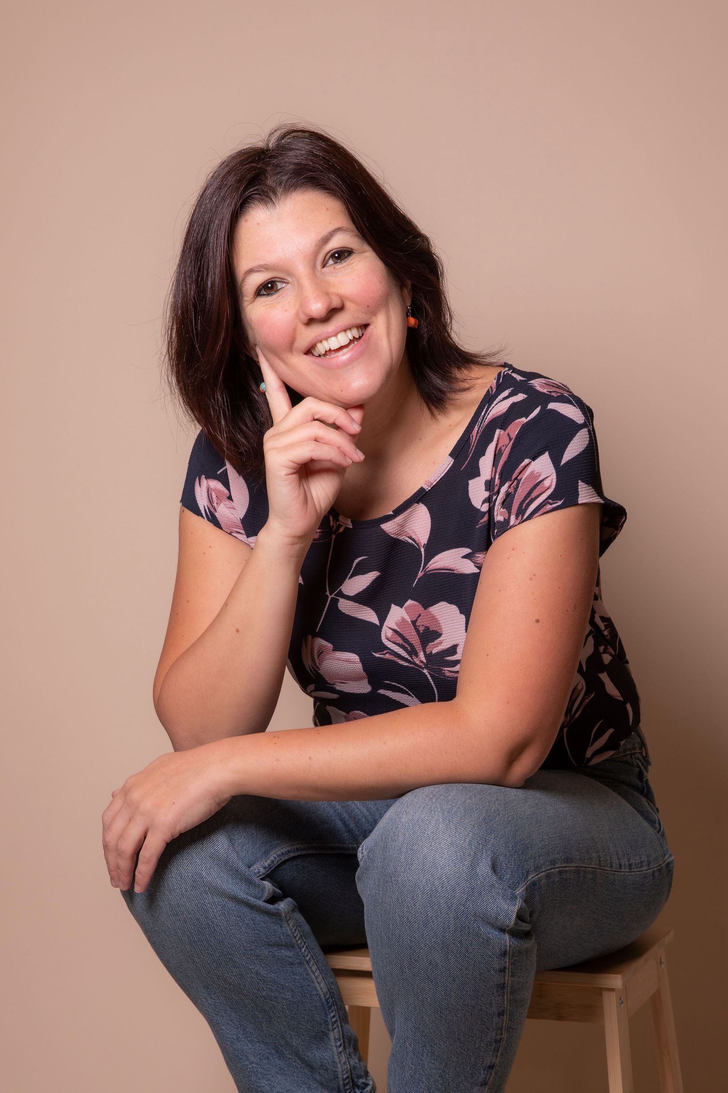 A person in a floral top and jeans sits casually on a chair against a neutral backdrop.