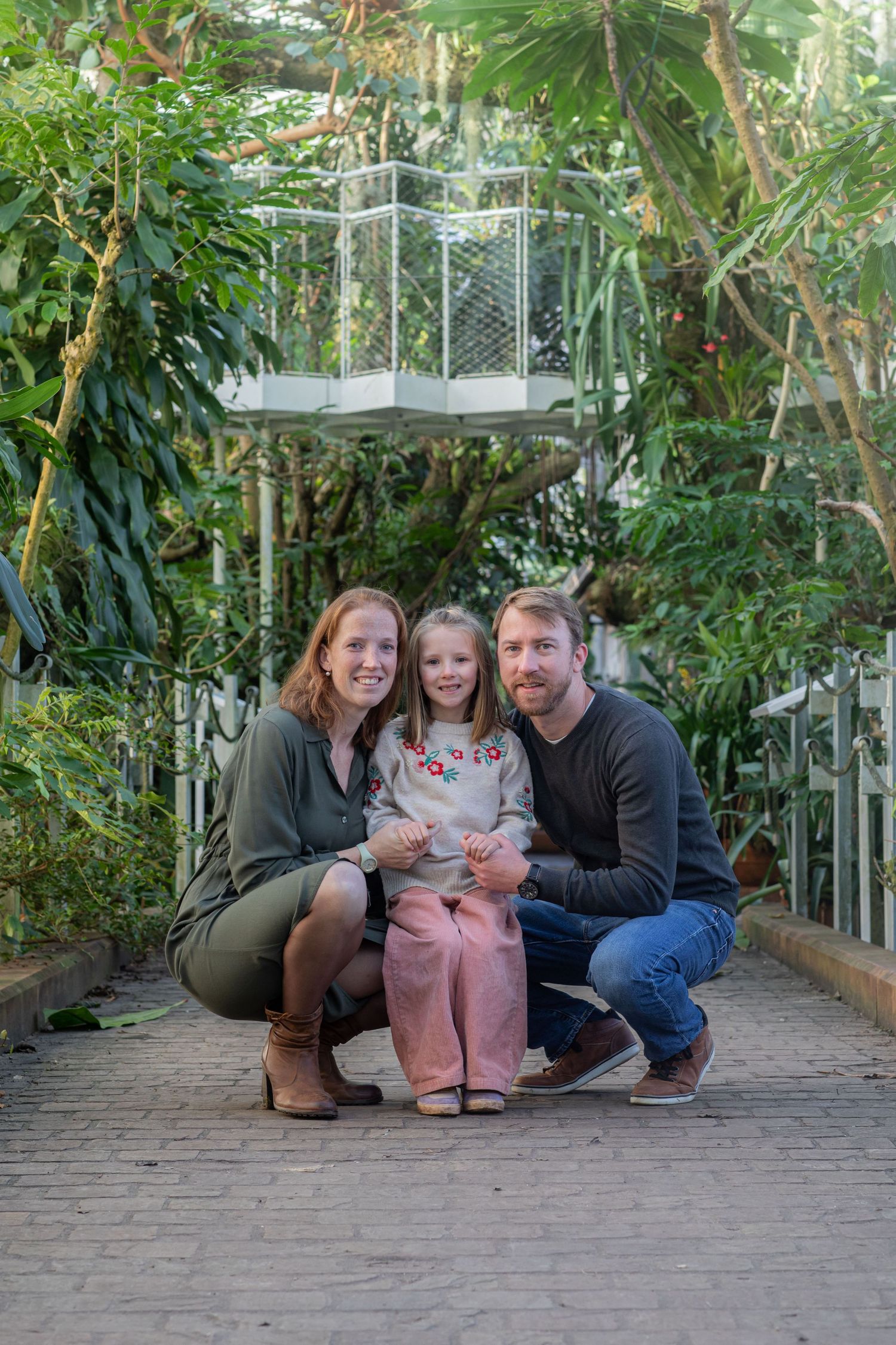 Family poses together on brick pathway surrounded by lush tropical greenery in outdoor garden setting.