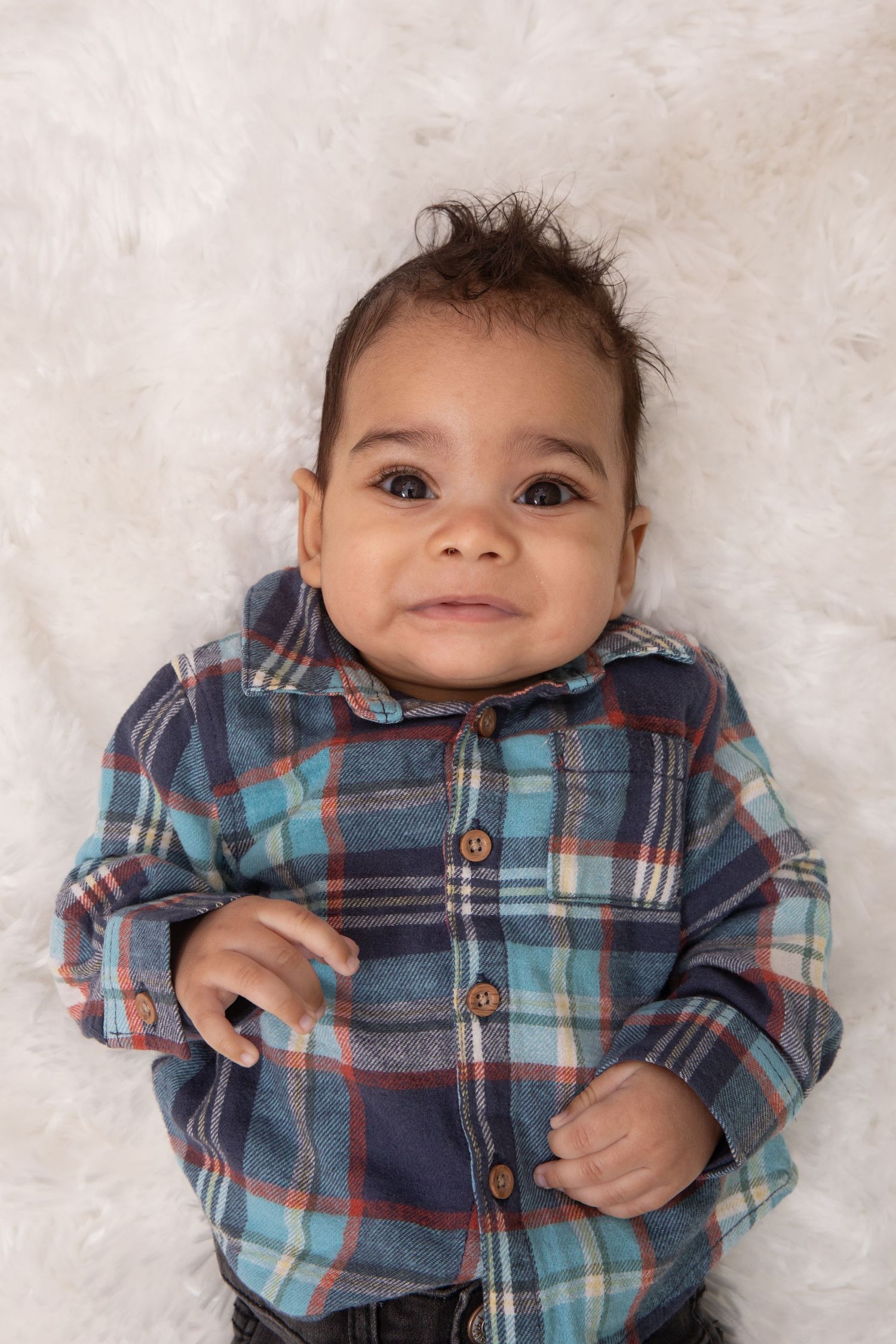Adorable baby boy in a plaid flannel shirt lying on a white fluffy blanket, smiling at the camera.