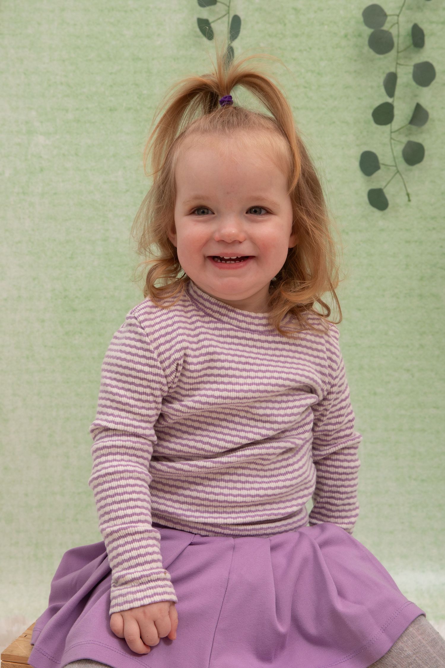 A smiling toddler girl with a ponytail wearing a striped top and purple skirt sits in front of a green backdrop.