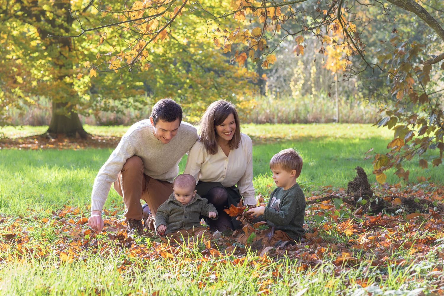 Family of four enjoying autumn leaves in a sunny park, with two young children sitting on grass surrounded by colorful foliage.