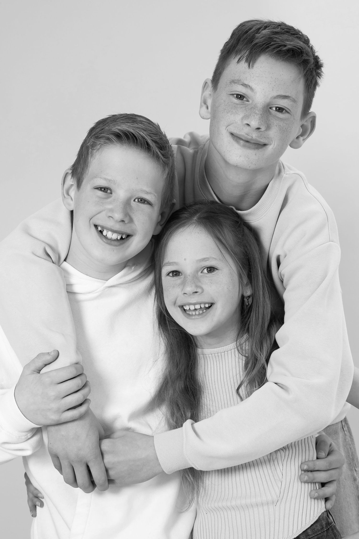 Black and white portrait of three smiling siblings hugging together, two boys and a girl, in casual white tops.