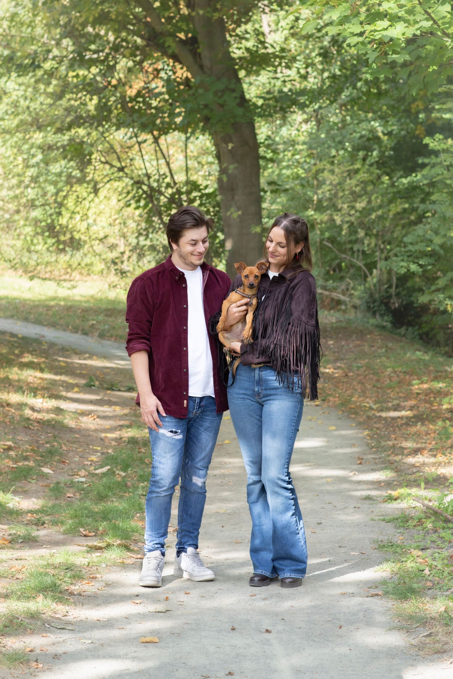 Couple walking together on a tree-lined path in autumn wearing burgundy and denim.
