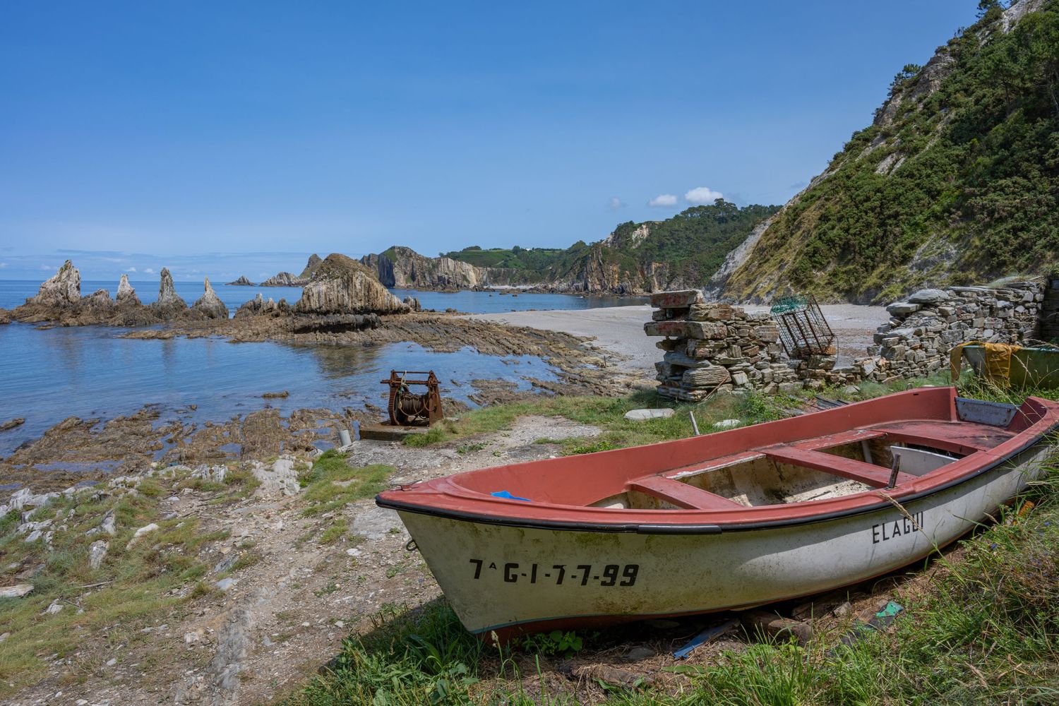 A weathered red and white wooden boat rests on a rocky shoreline beside a calm coastal bay with dramatic rock formations.