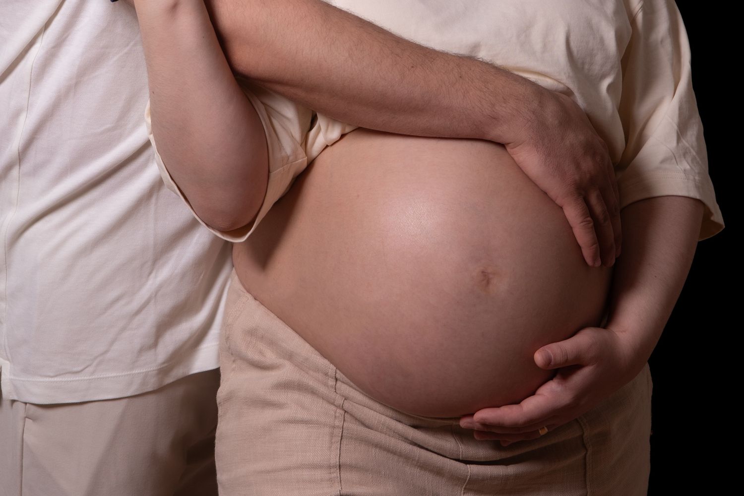 Pregnant woman with partner's hands gently cradling her bare baby bump against a dark background.