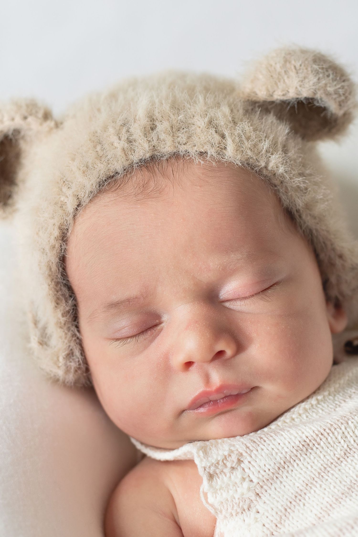 Sleeping newborn baby wearing a soft cream-colored knit hat on white bedding.