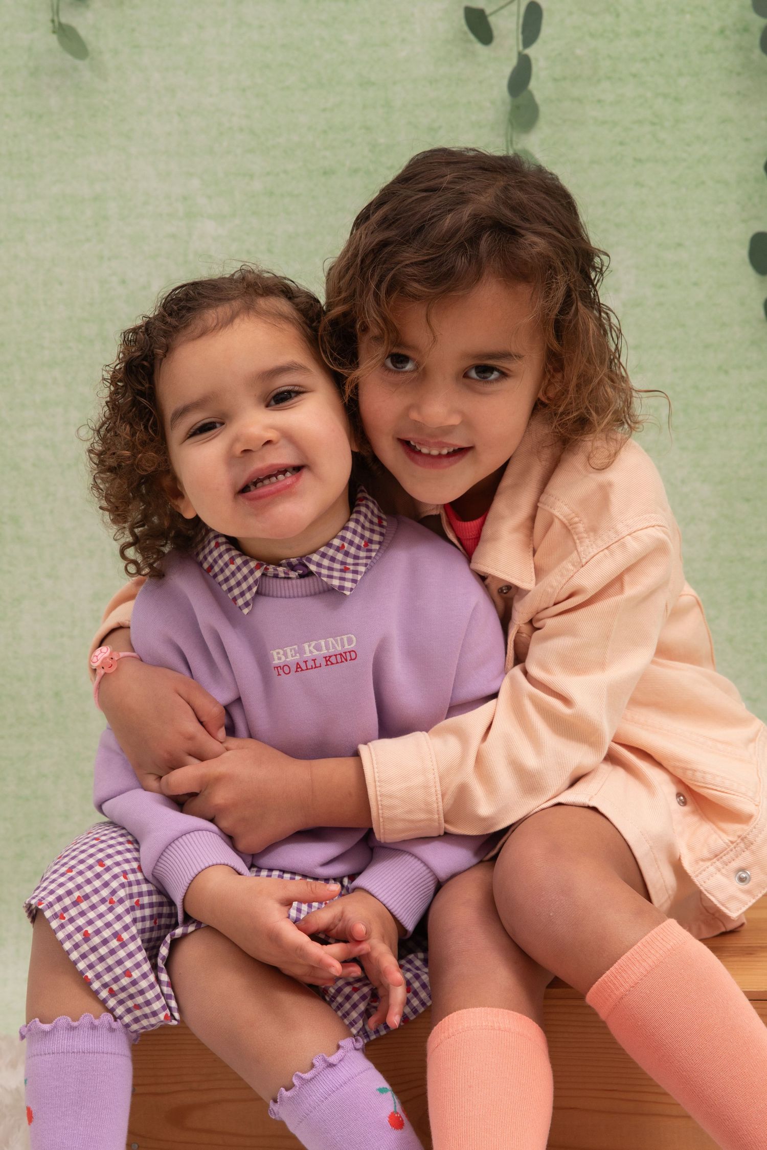 Two young girls with curly hair smiling and hugging, wearing purple and peach outfits with gingham accents.