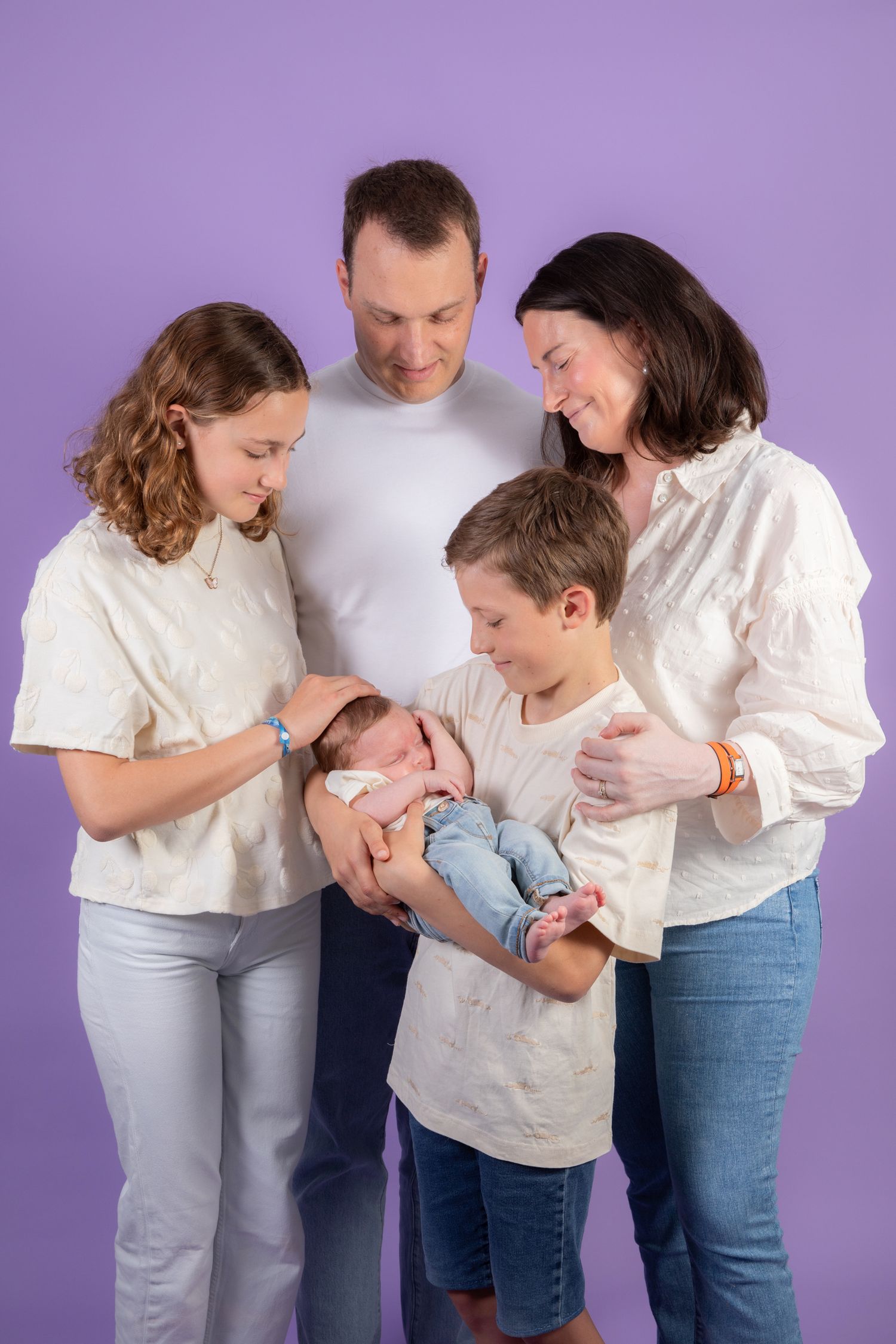 Family gathered around newborn baby against purple background wearing coordinated white and denim outfits.