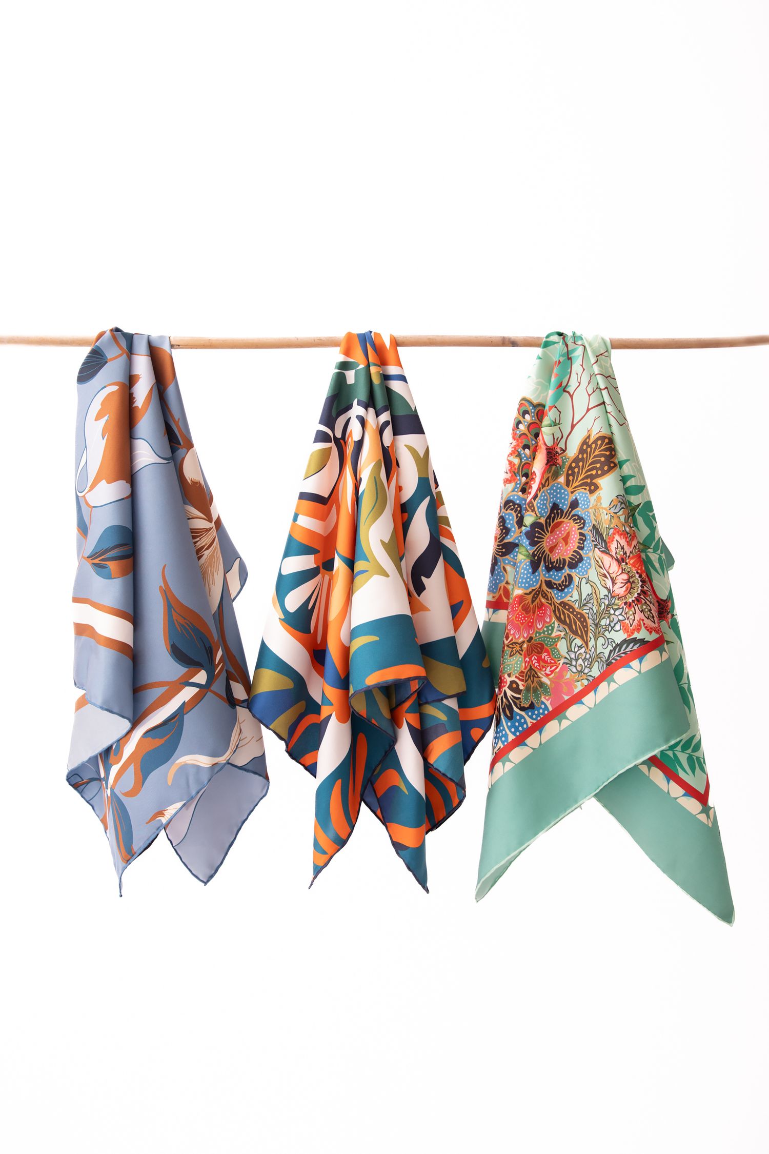 Colorful silk scarves with floral and geometric patterns hanging on a copper rod against a white background.