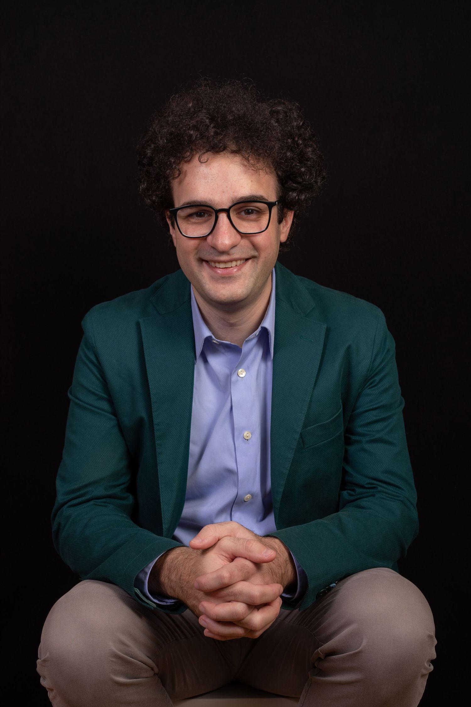 Person in green blazer and purple shirt seated against black background.