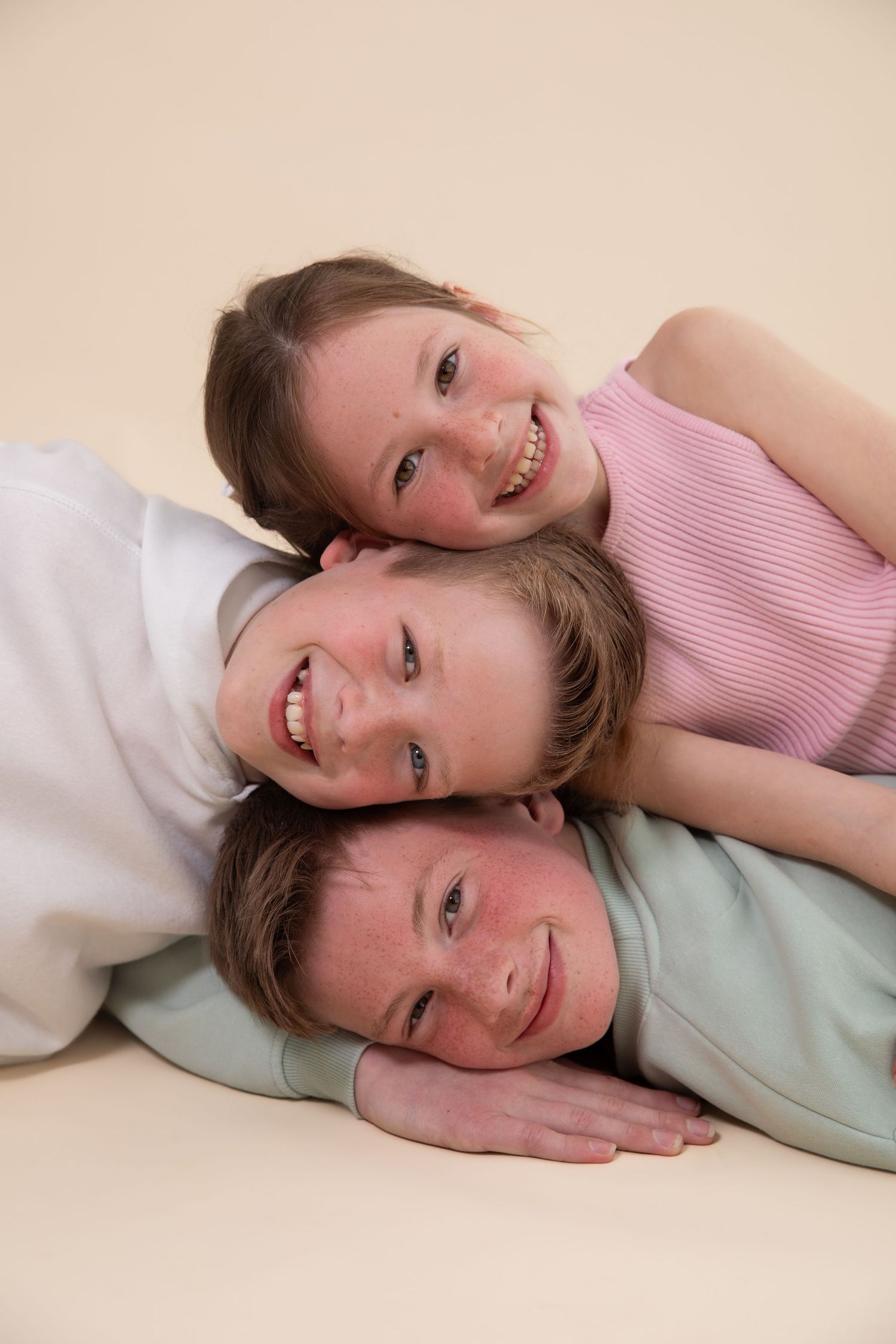 Three smiling children stacked together lying down, posing for a fun portrait photo against a beige background.