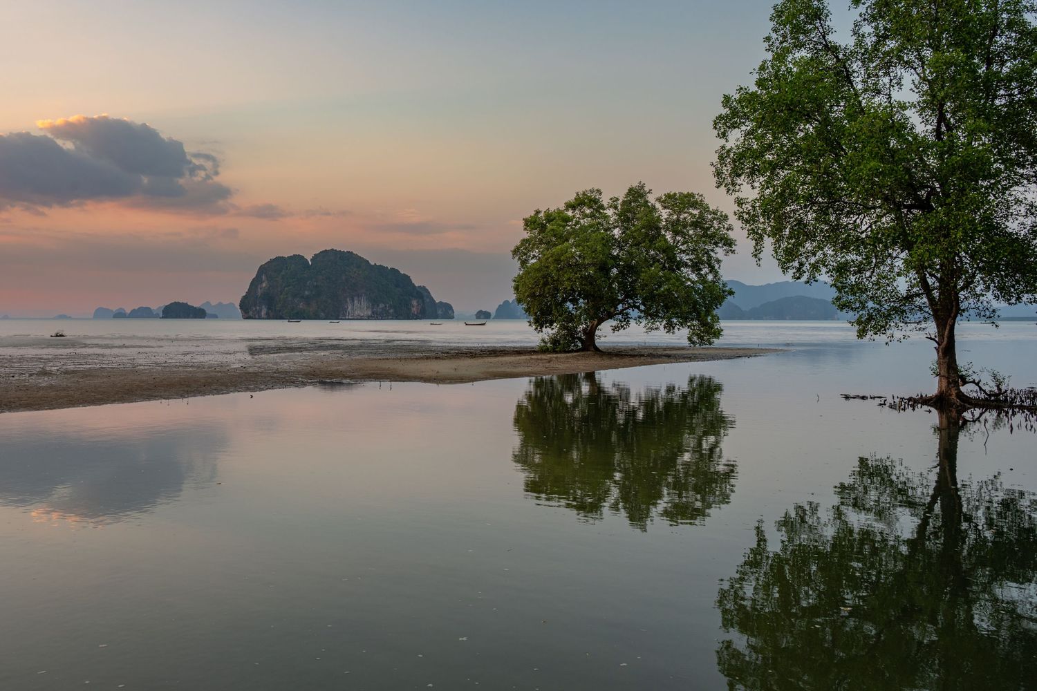 Serene tropical bay at sunset with limestone karst islands, reflective calm water, and lush green trees mirrored on the surface.