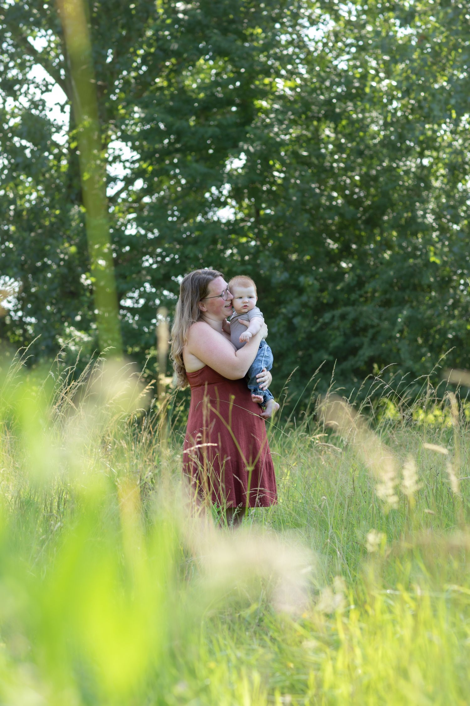 A mother in a red dress holds her baby in a lush green meadow surrounded by tall grass and trees.