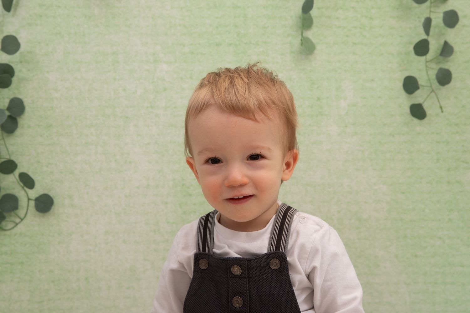 Smiling toddler boy with blonde hair wearing dark overalls over white shirt, posed against green botanical backdrop.