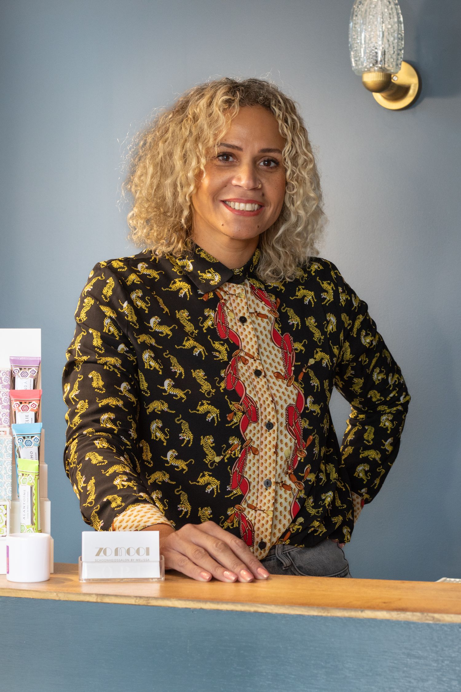 A person with curly blonde hair wearing a black and yellow patterned top sits at a desk with skincare products.