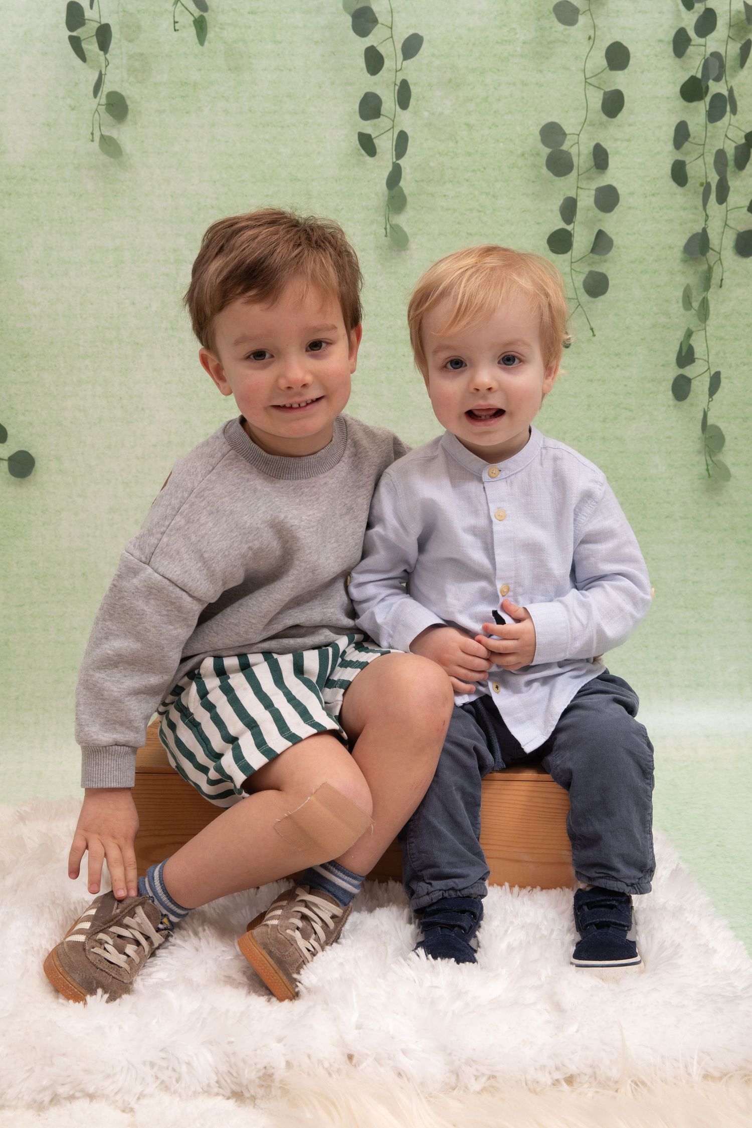 Two young boys sitting together on a pot in front of a green botanical backdrop on a white fluffy rug.