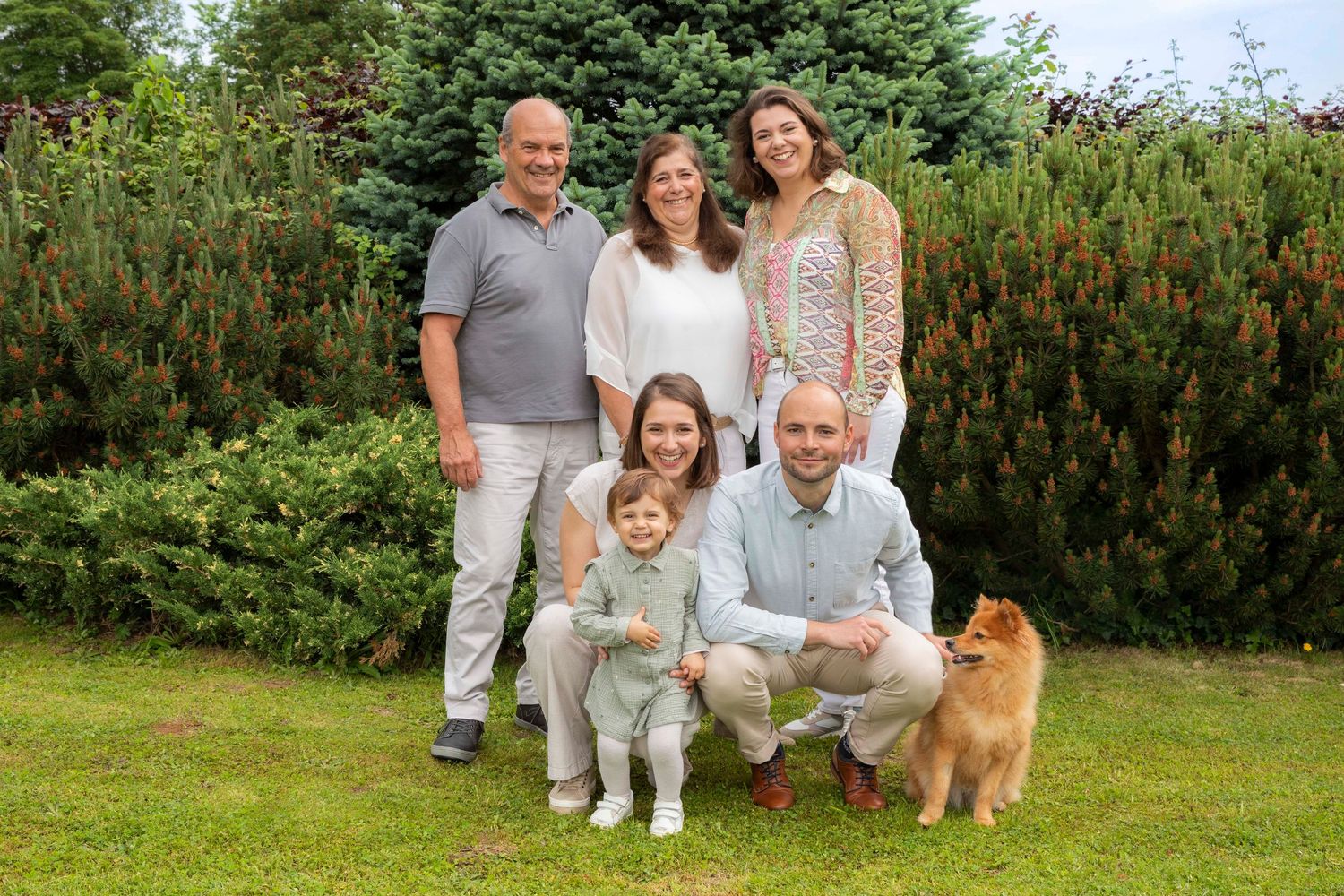 Family of six with a Pomeranian dog posing outdoors on green grass with lush shrubs in the background.