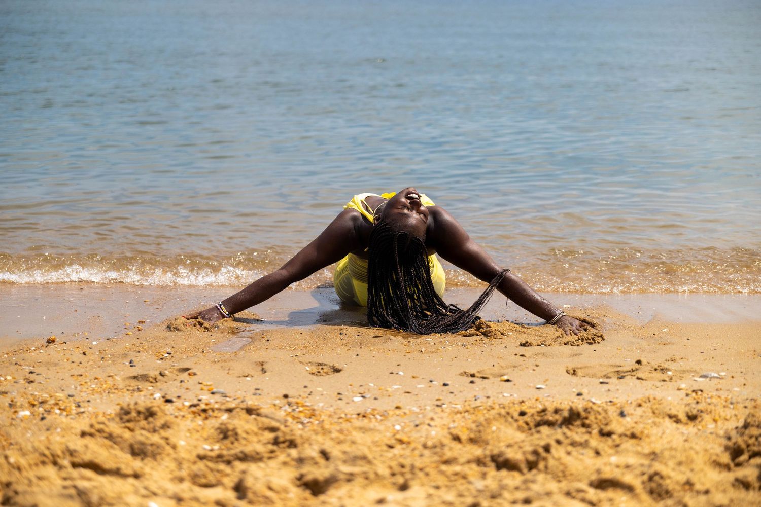 Woman in yellow swimsuit lying on sandy beach with arms spread, relaxing by calm blue ocean waters.
