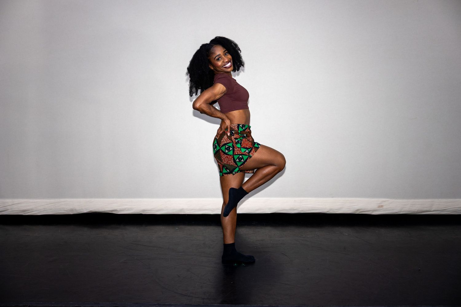 Black woman in African print skirt poses with one leg raised on a dark performance stage.