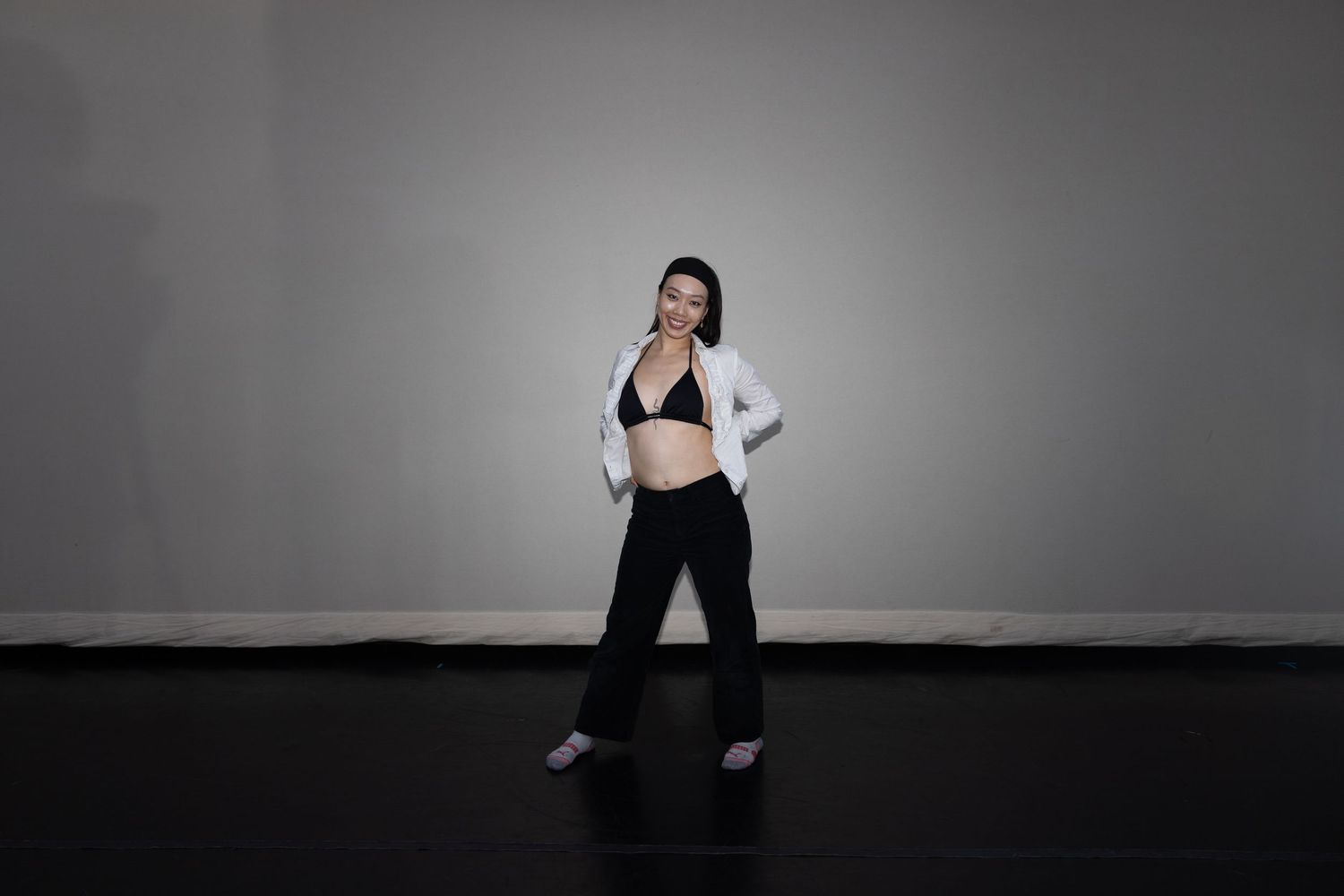 Woman in black bralette and pants with open white blazer posing in dance studio.