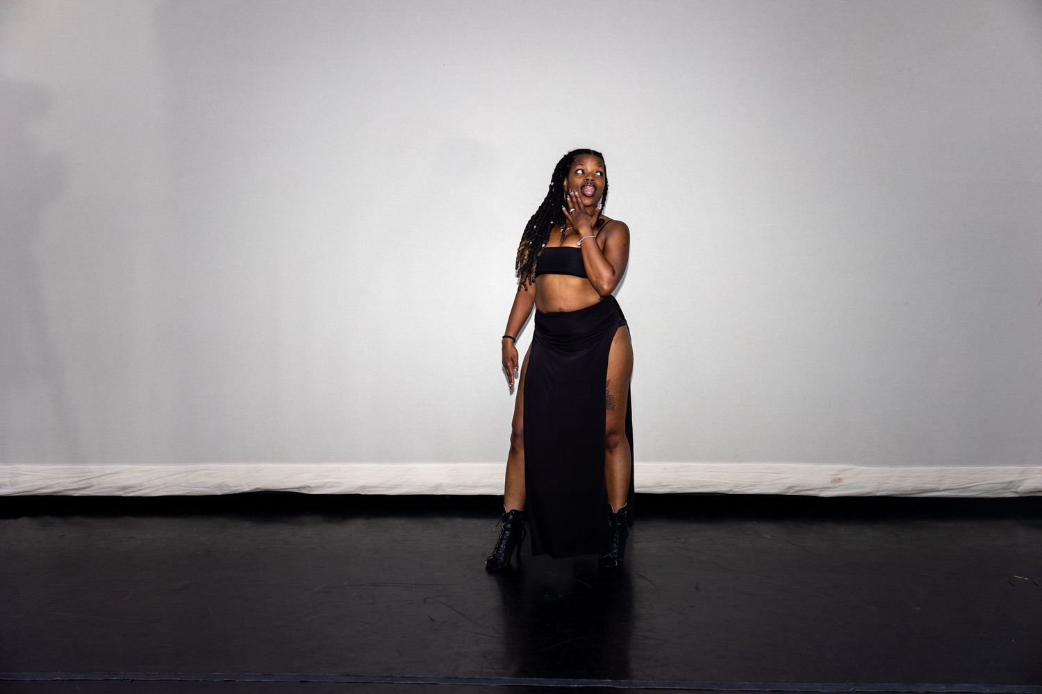 Elegant woman in black two-piece outfit with high-slit skirt poses on dark stage floor.