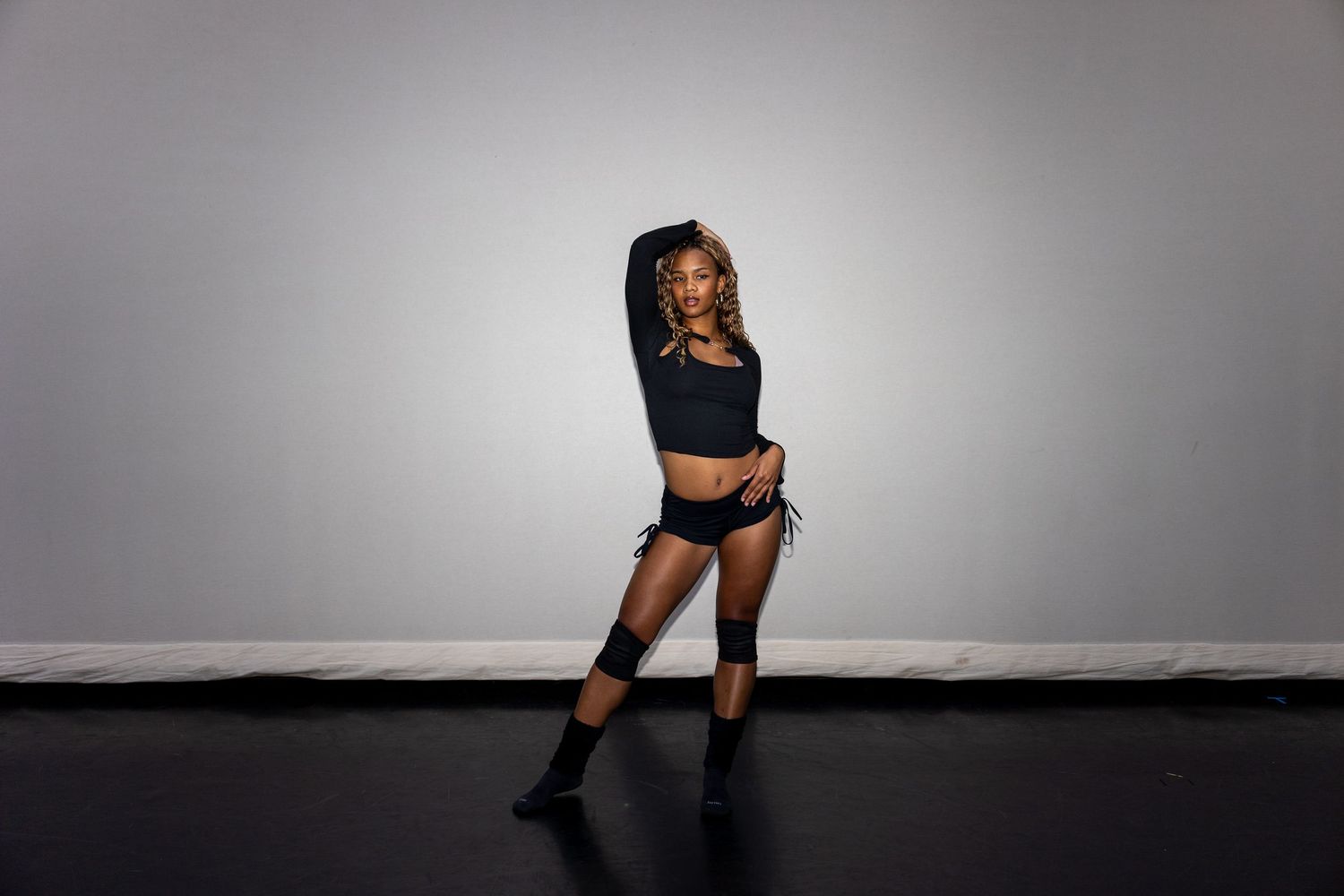 A dancer in black crop top, shorts, and knee-high boots poses dynamically against a gray studio background.