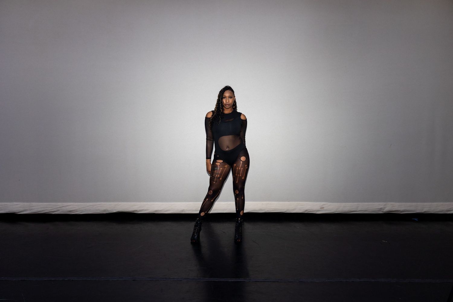 A dancer in a black mesh bodysuit and heels strikes a bold pose in a professional gray studio.